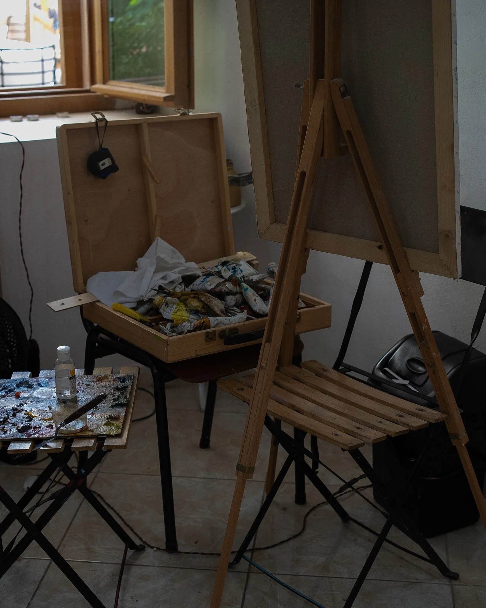 An art studio with an easel, a wooden box filled with paint tubes, a small table with an artist's palette and a bottle of paint, and a window in the background.