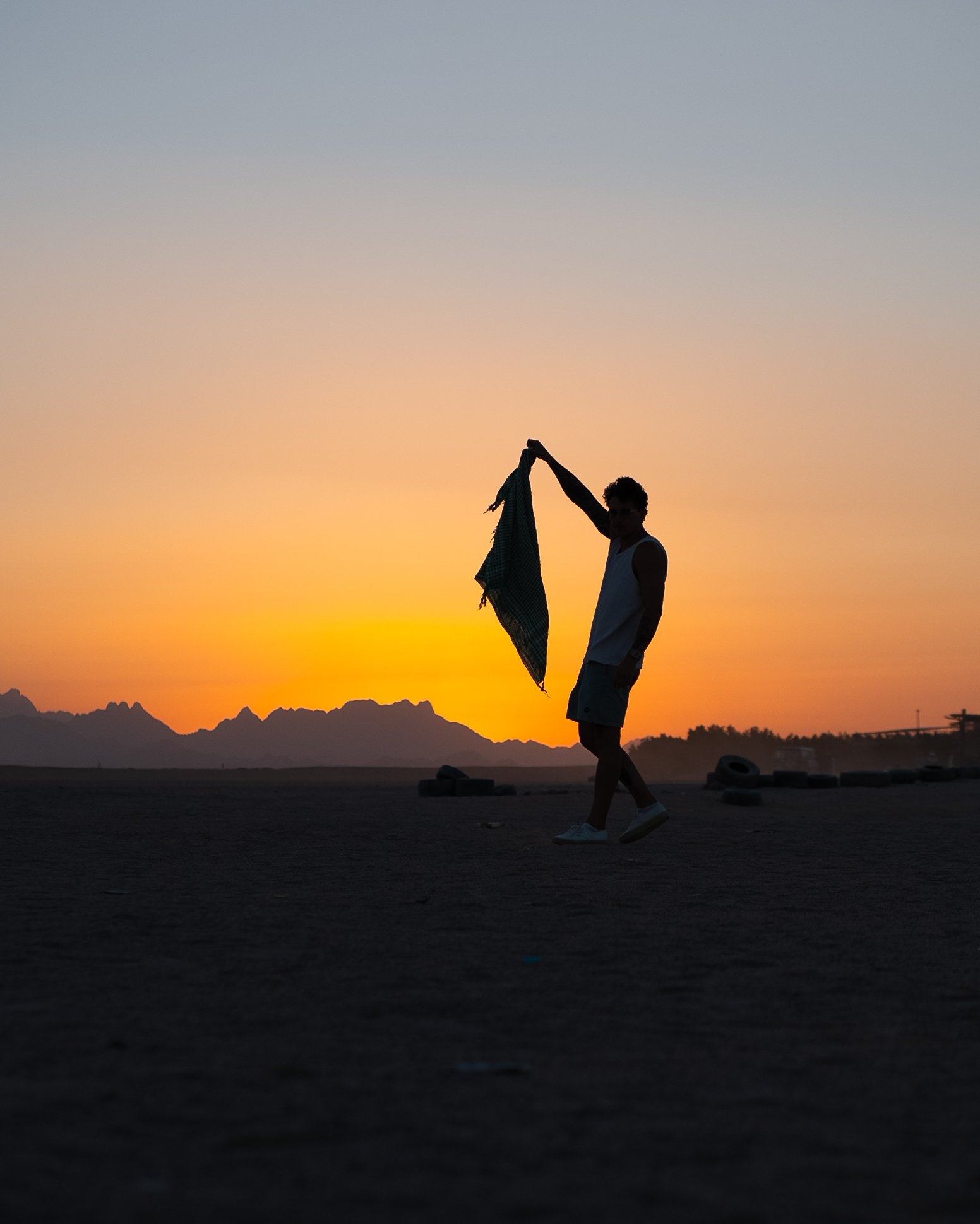 Silhouette of Bogdan Kosinov walking on a desert at sunset, holding a scarf in one hand, with mountains and tires in the background.