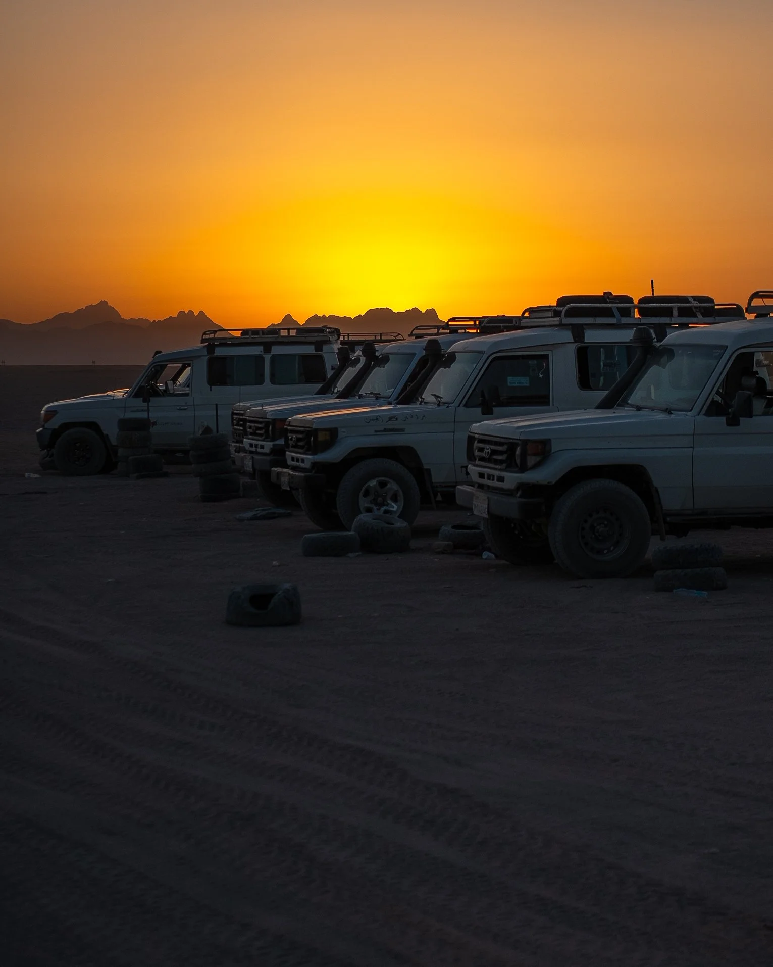 Several white off-road vehicles parked on a dirt terrain during sunset with mountains in the background.