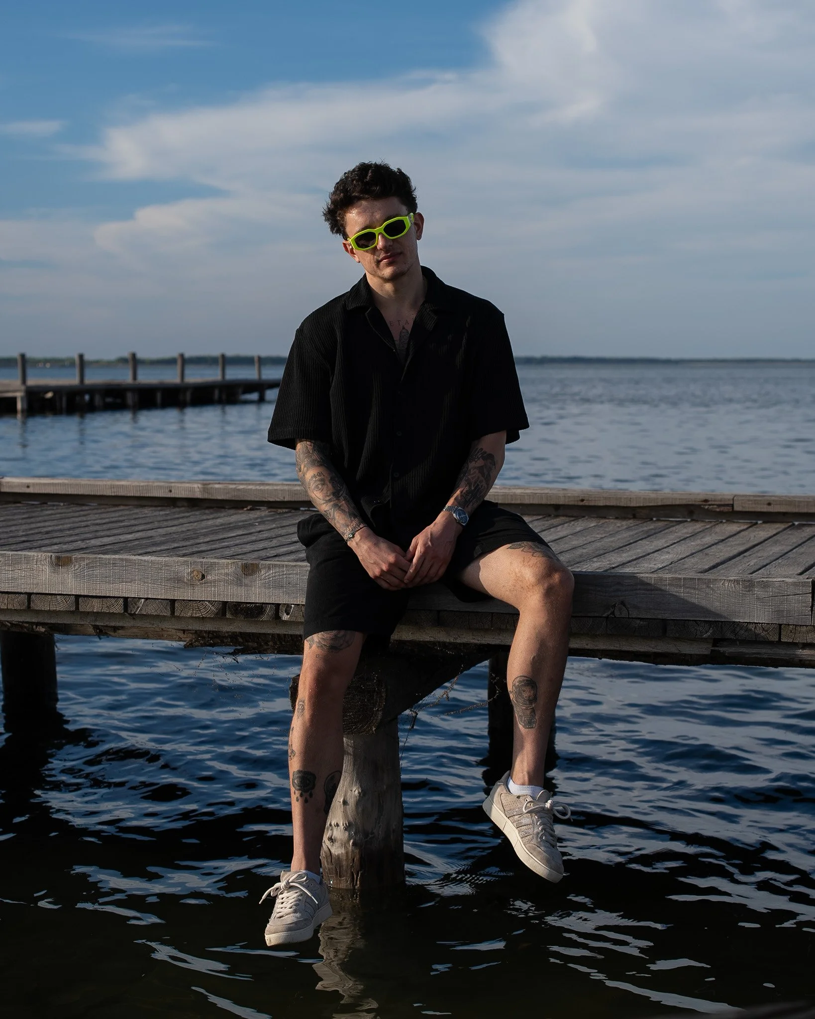 IMBK sitting on a wooden dock by the water, wearing a black shirt, black shorts, white sneakers, yellow sunglasses, and a wristwatch, with a background of a lake and a cloudy sky.
