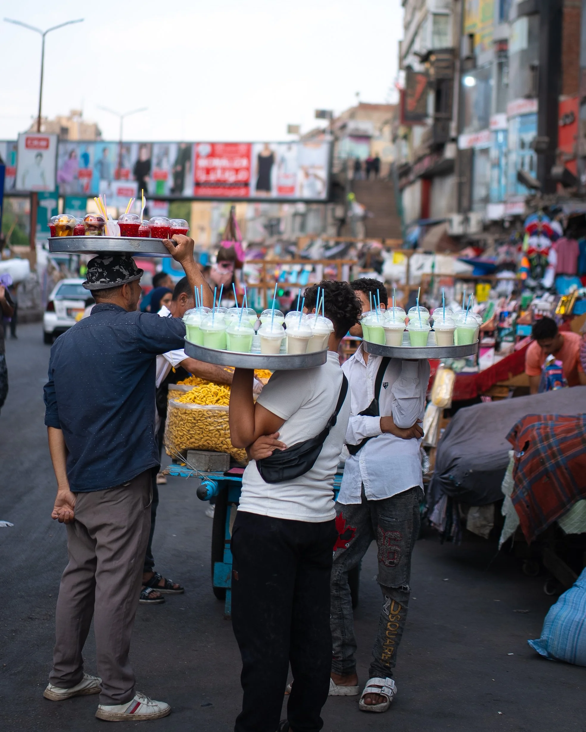 Street scene with vendors selling colorful drinks on trays, surrounded by people, market stalls, and buildings.