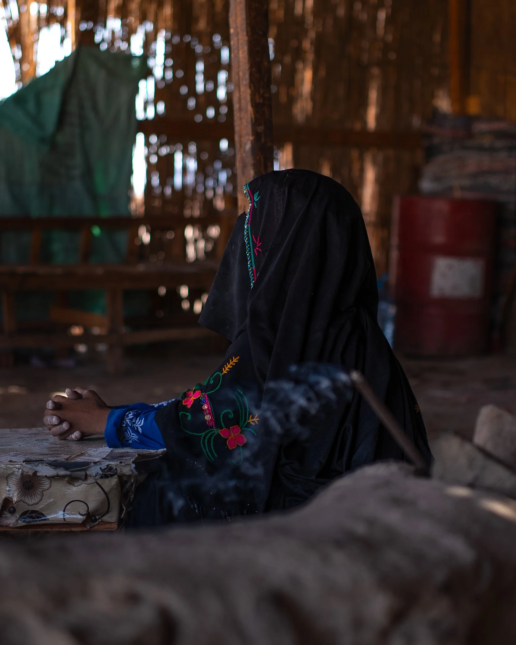 A woman in traditional embroidered clothing sitting in a rustic, wooden hut with her hands folded on a table.