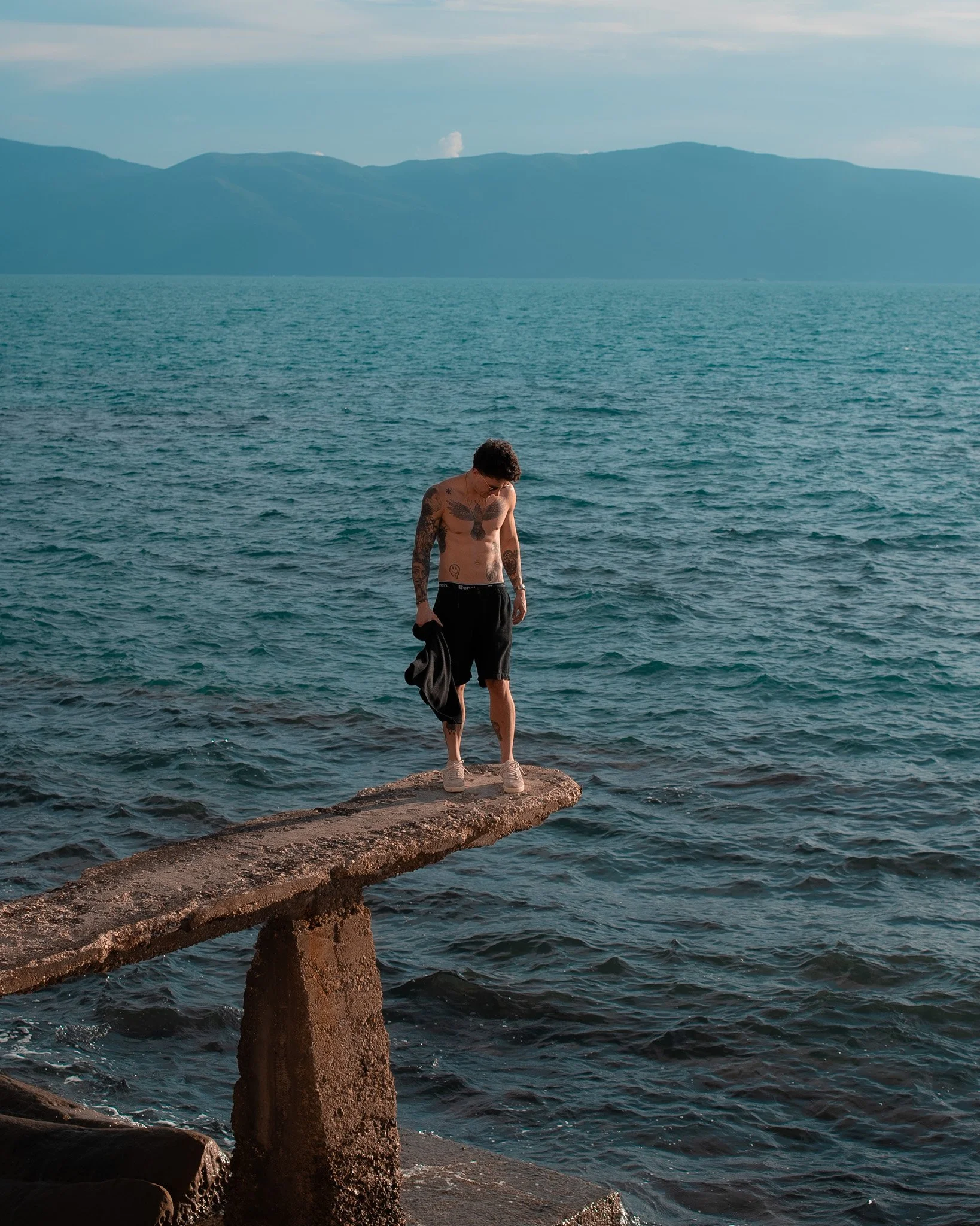 A shirtless man with tattoos, wearing shorts and sneakers, stands on a narrow, weathered concrete pier extending over water, with mountains in the background.