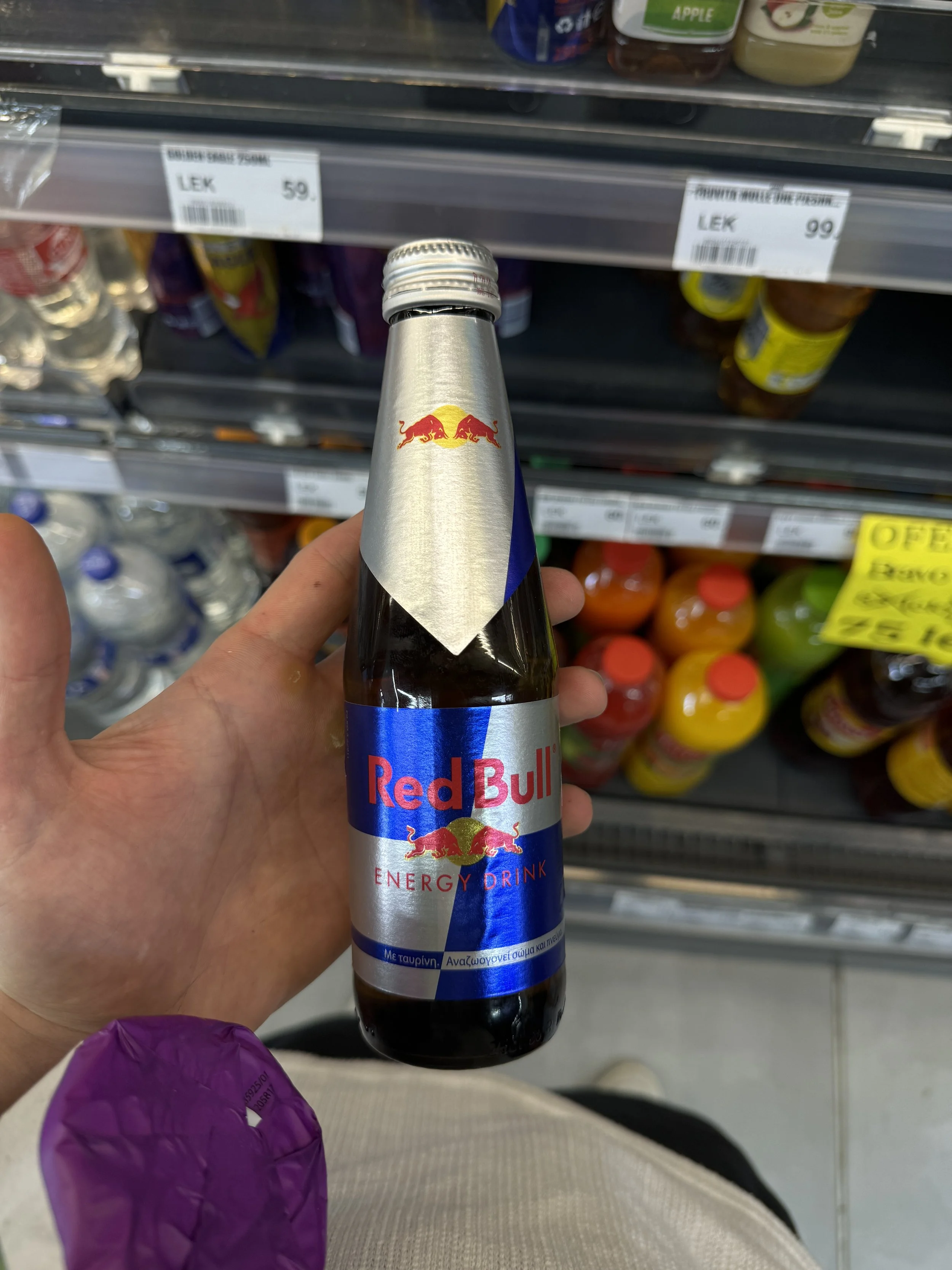 A person holding a small bottle of Red Bull energy drink in a grocery store aisle with shelves of bottled beverages in the background.