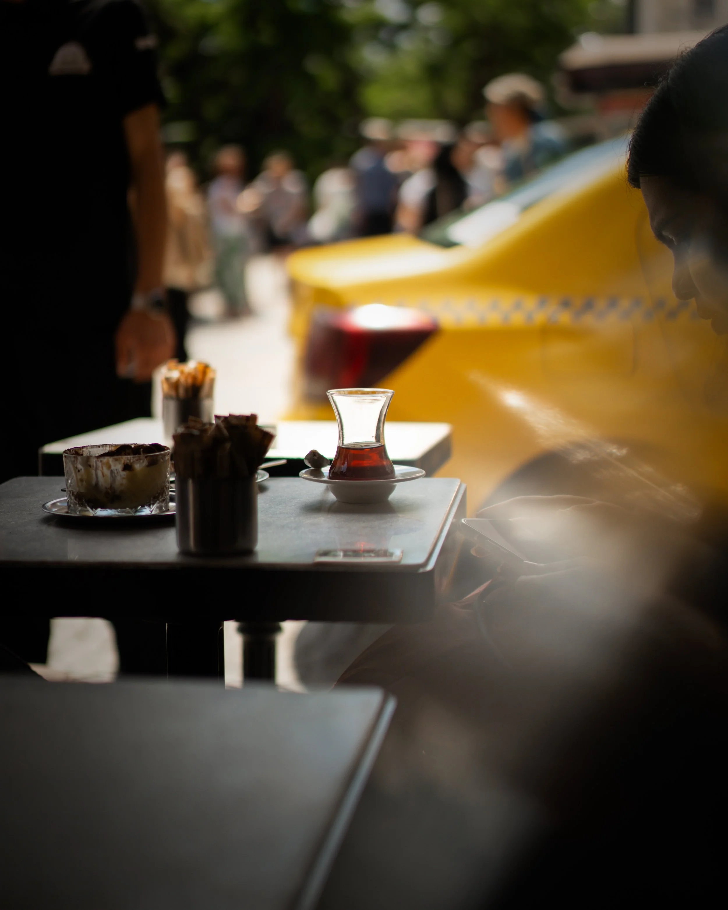 A table with a Turkish tea glass filled with tea, a plate of desserts, and a container holding toothpicks, set outside with blurred people and cars in the background.