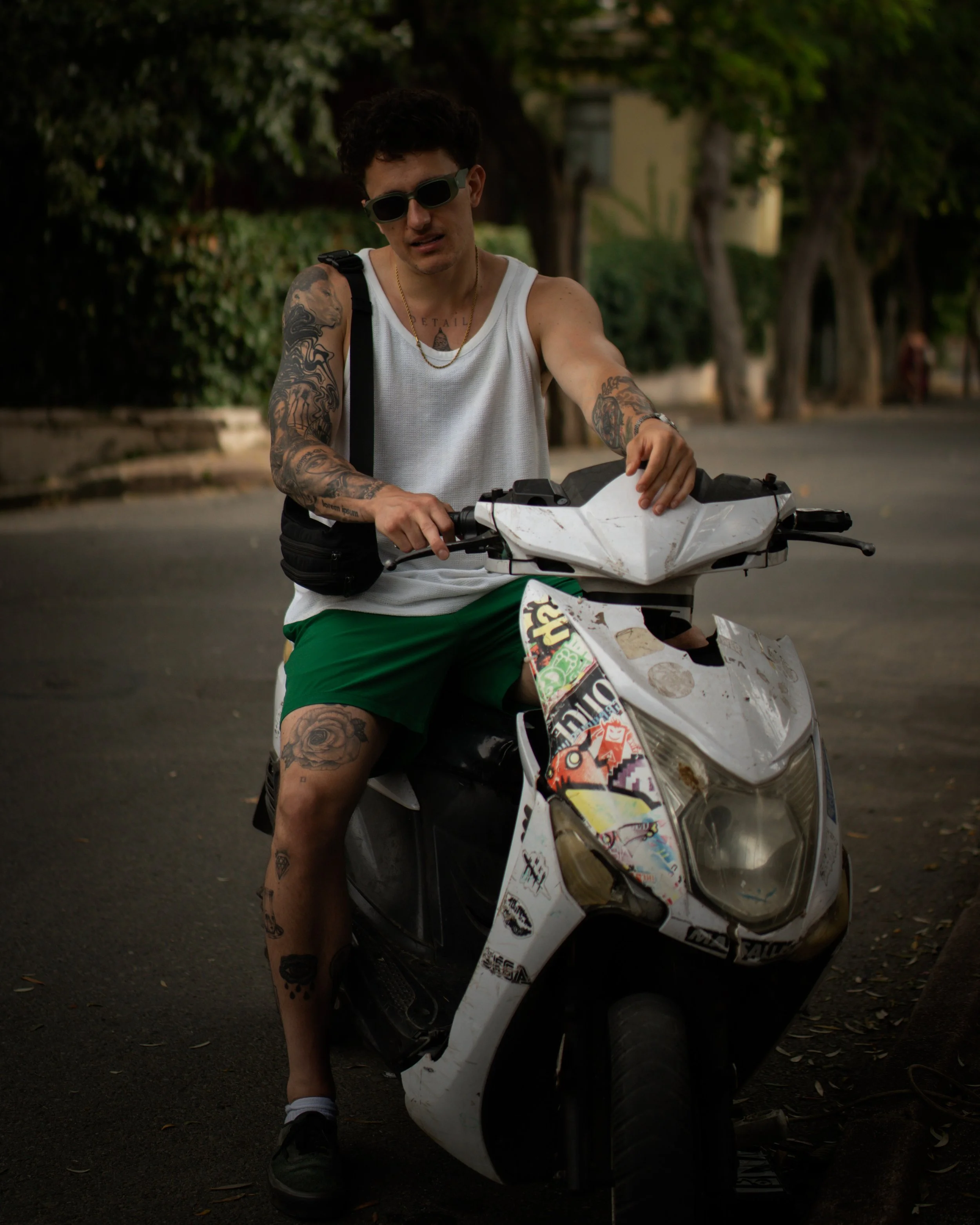 IMBK wearing sunglasses, a white tank top, and green shorts, sits on a white scooter parked on a street with trees in the background.