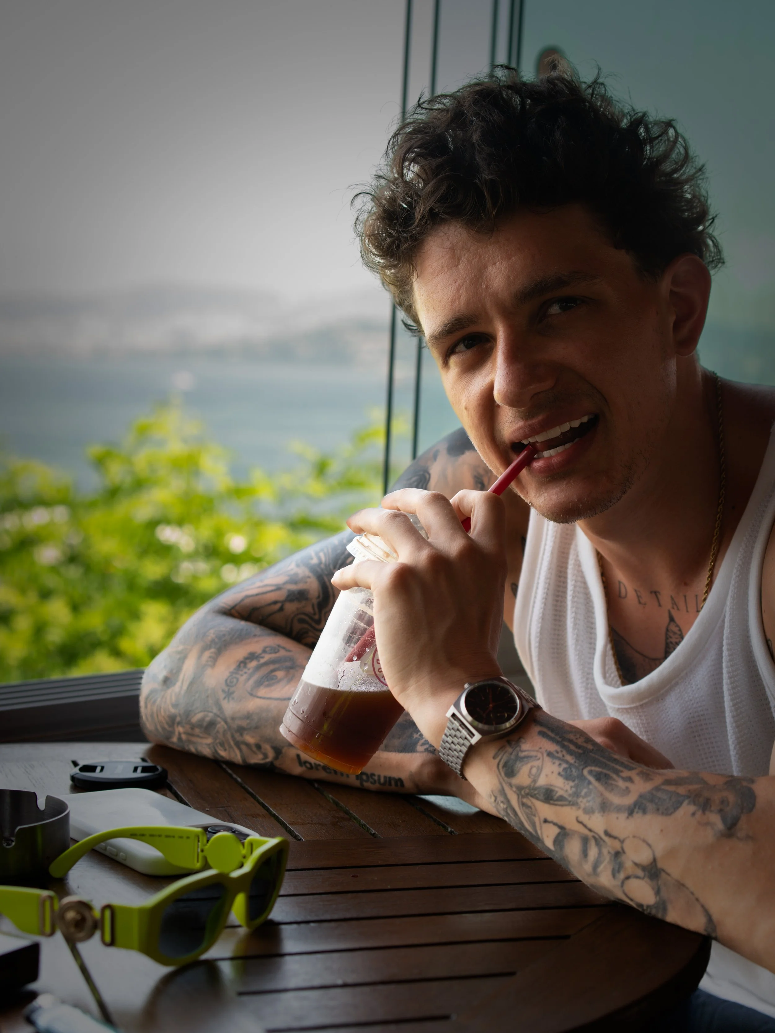 Bogdan Kosinov, wearing a white sleeveless shirt and a watch, sitting at a wooden table with sunglasses, a phone, and a drink, smiling and looking at the camera, with a scenic outdoor view of greenery and water in the background.