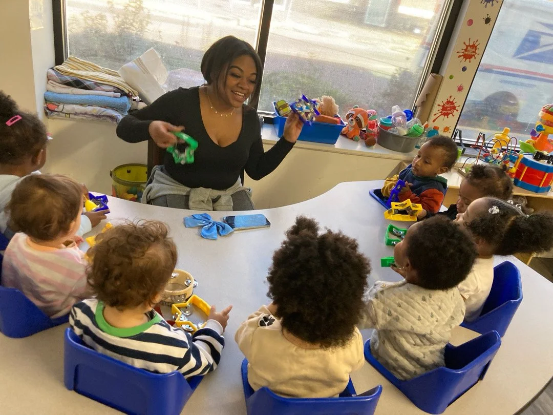 Teacher leads a music activity with toddlers seated around a table. Children hold colorful instruments and watch as she demonstrates how to play them.