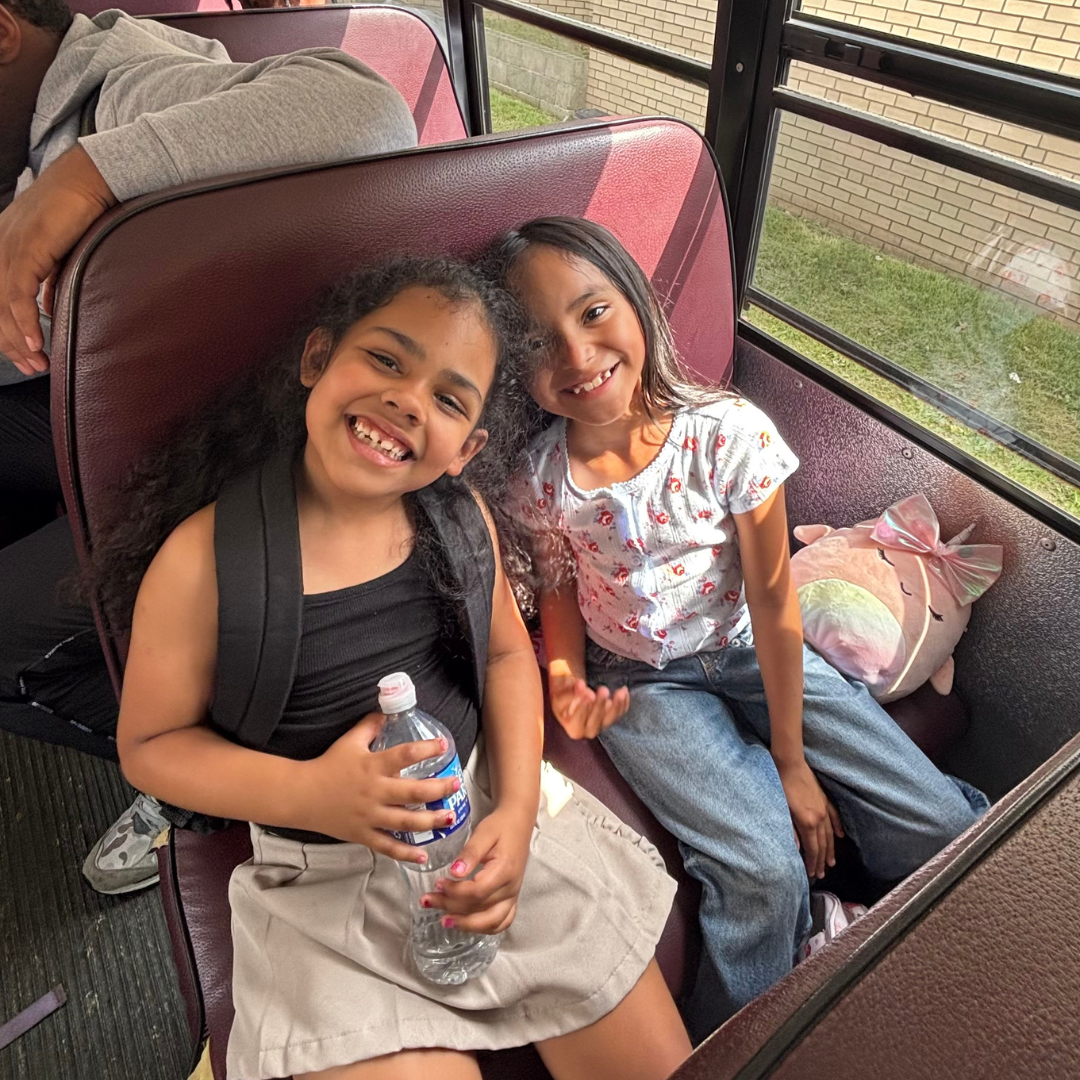 Two young girls seated on a bus seat, smiling and enjoying their time. One girl is holding a water bottle, and there is a unicorn plush pillow beside them. The bus window shows a grassy area and a brick wall outside.
