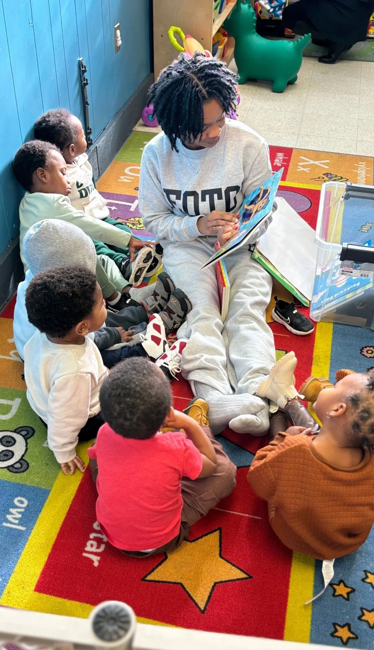 Teacher sits on a colorful classroom rug reading a book to toddlers gathered around her. The children listen closely during storytime.