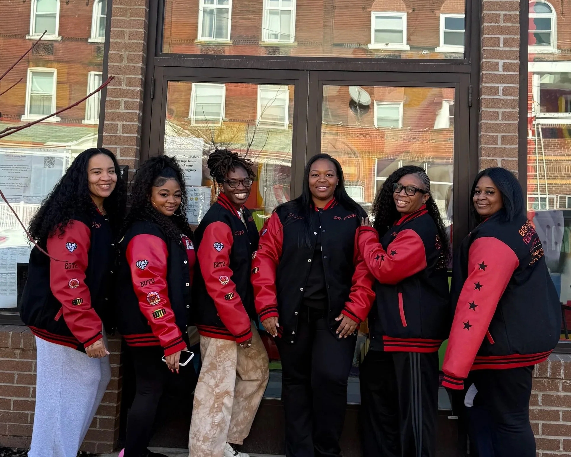 Group picture of the administration team wearing black and red matching letterman jackets with EOTO patches and custom names. The group of women are all smiling in a line with Dr. Dom in the center.