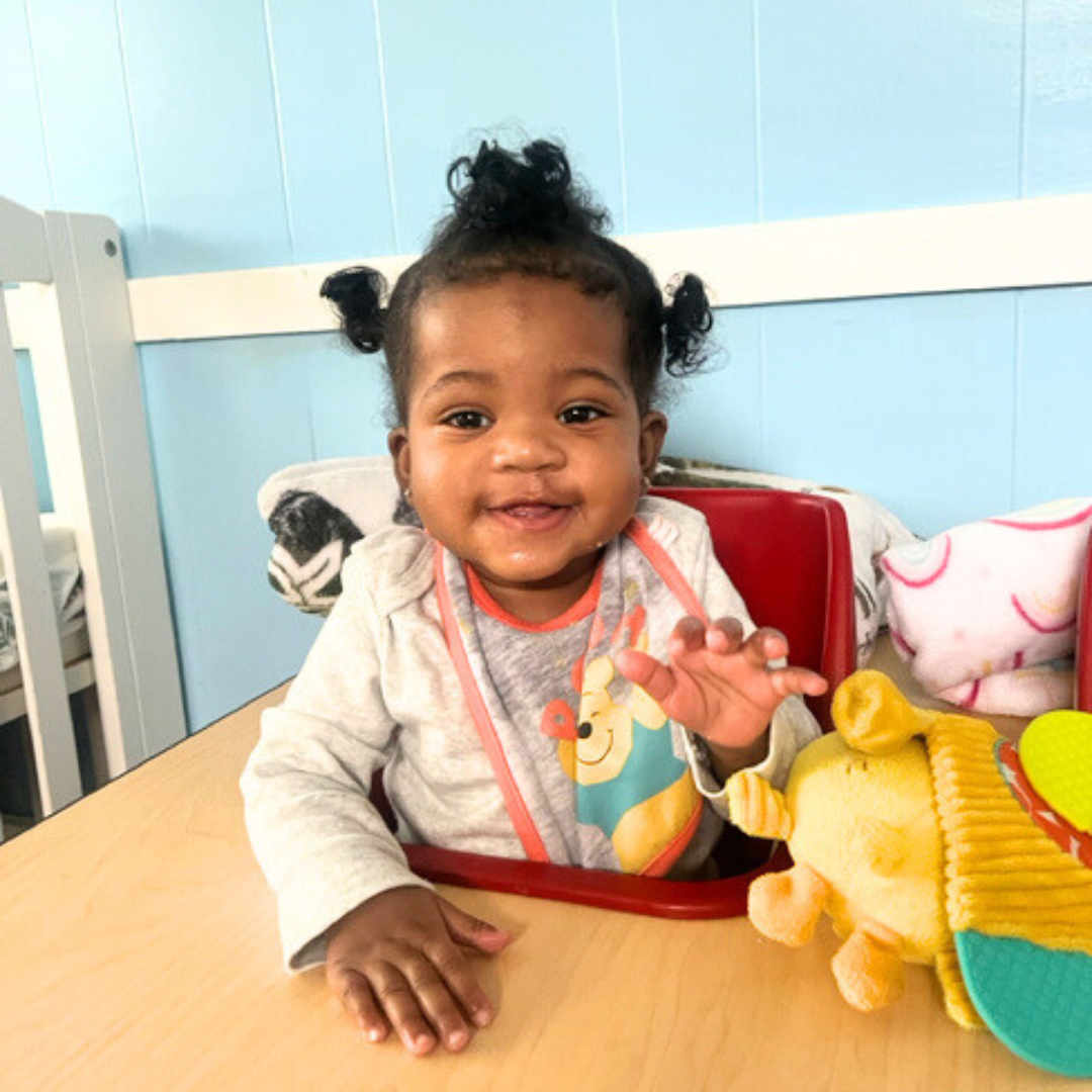 Smiling infant girl sitting in a red seat at a classroom table, wearing a gray outfit with a colorful bib and playing beside a yellow plush toy in a safe, comfortable classroom environment.