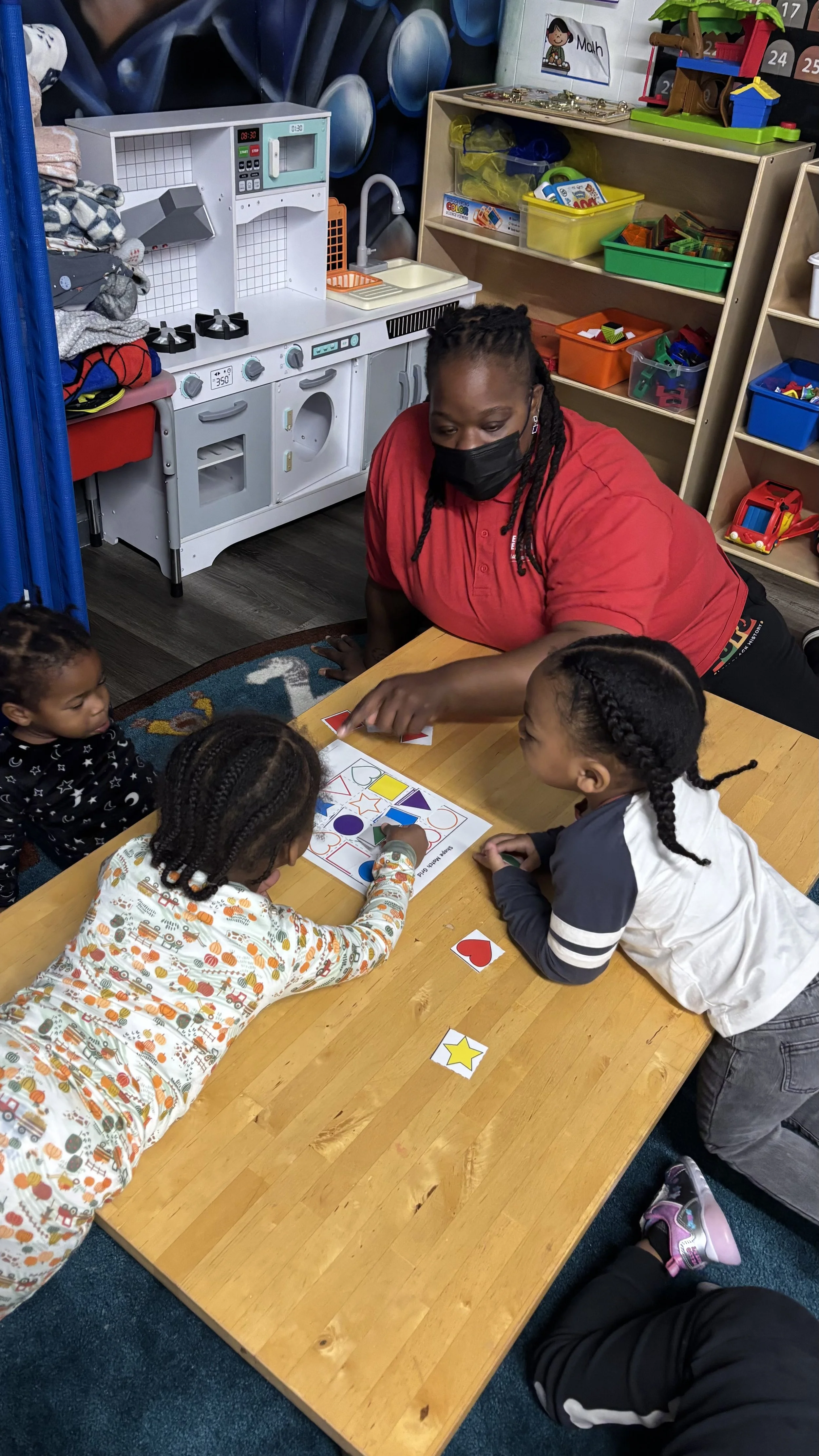 Teacher sits at a table guiding toddlers through a shapes matching activity. Children focus on placing colorful shape cards onto a worksheet in the classroom.
