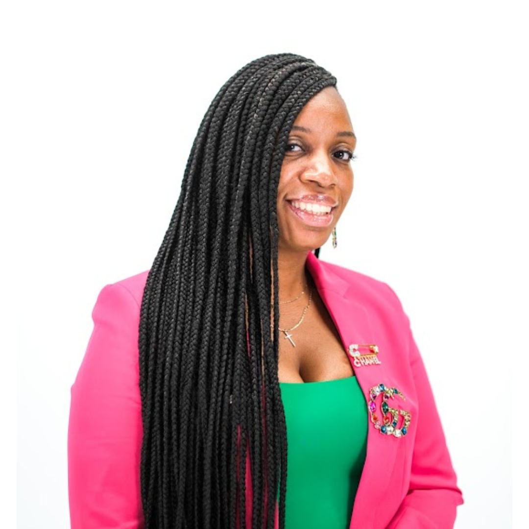 Woman with long black braids smiling, wearing a pink blazer with a prominent pin, a green top, and jewelry, standing against a plain white background.