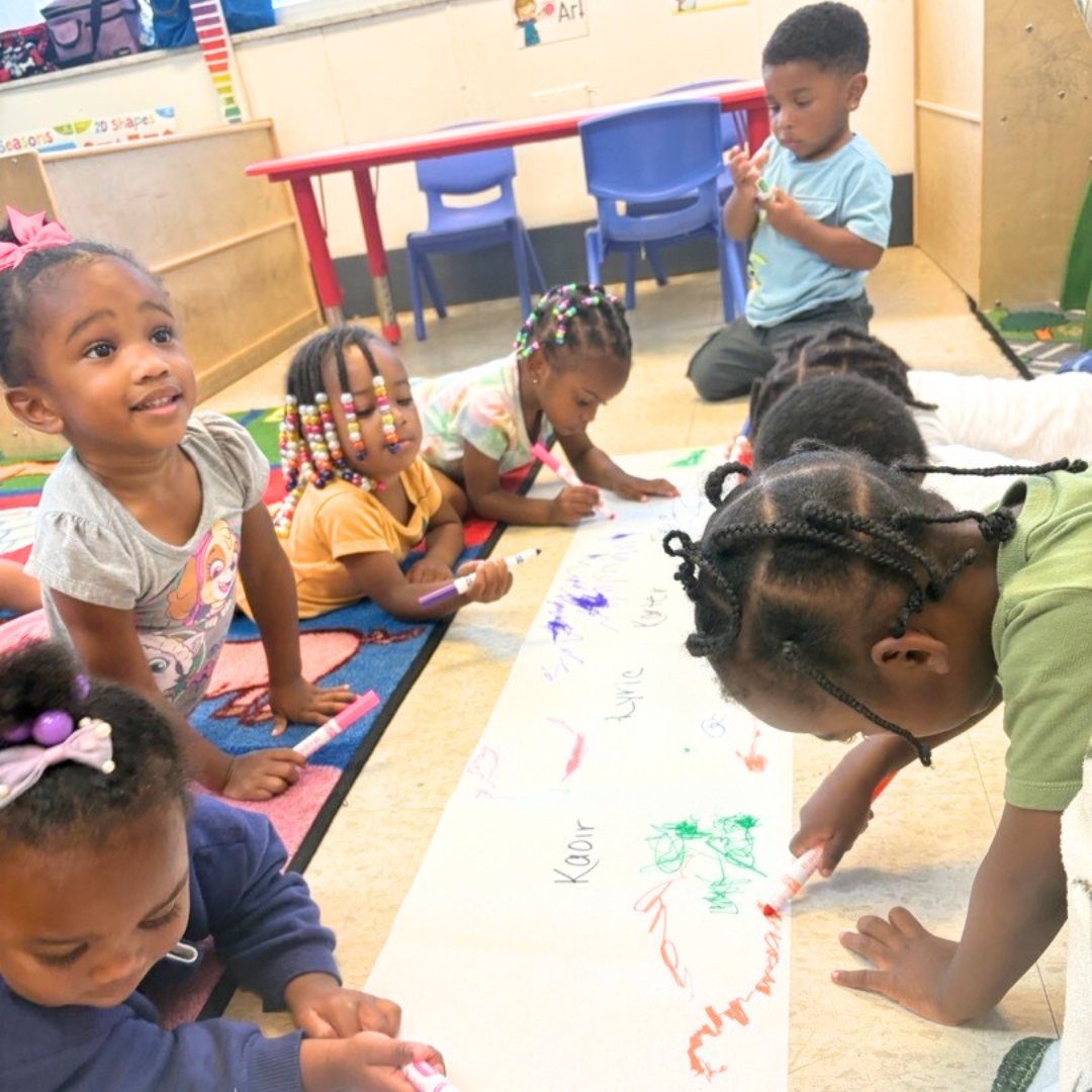 Toddlers sit on the classroom floor coloring together on a large sheet of paper. Children use markers to draw and write under their names during a group activity.