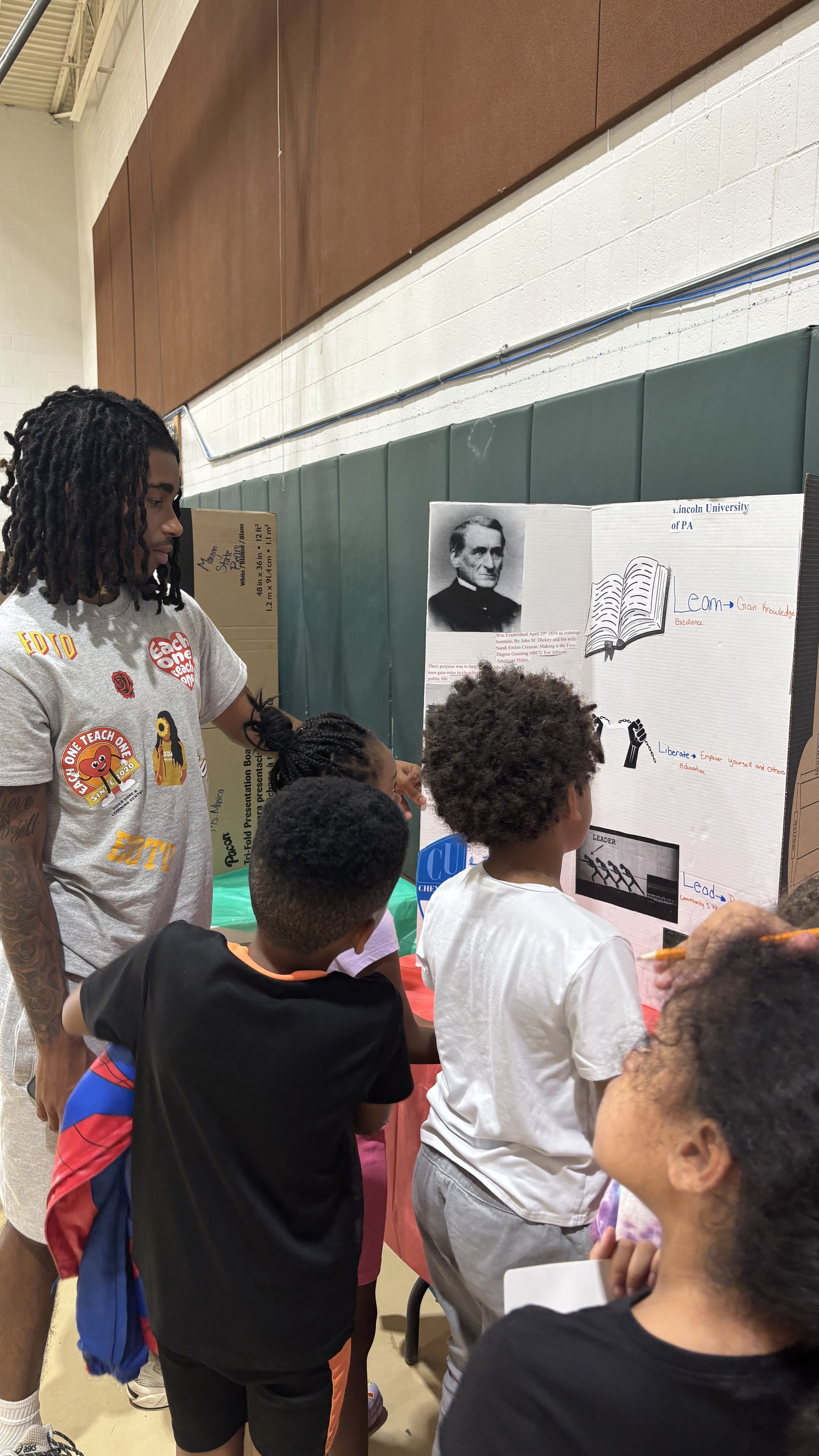 One Teach One’s Cultural Fair. The tri-fold display highlights the university’s history, values, and legacy as an HBCU.Children listen and engage in learning about culture, leadership, and education in a bright gym setting.
