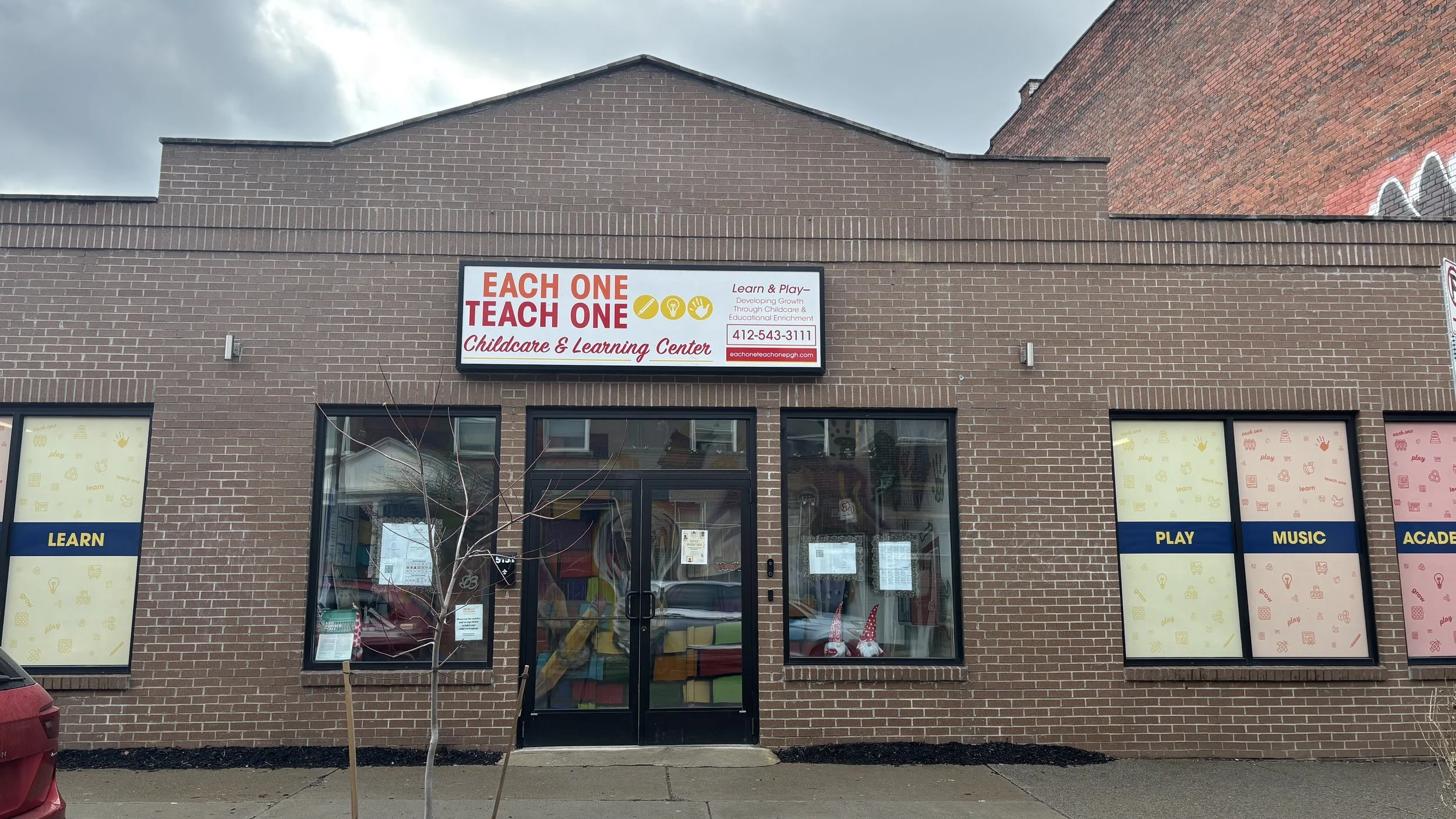 Facade of a childcare and learning center with a brick exterior, glass doors, and windows displaying colorful decorations. A sign above the entrance reads "EACH ONE TEACH ONE Childcare & Learning Center" with additional information and the phone number. The windows have words "LEARN," "PLAY," "MUSIC," and "ACADEMY" with relevant icons.