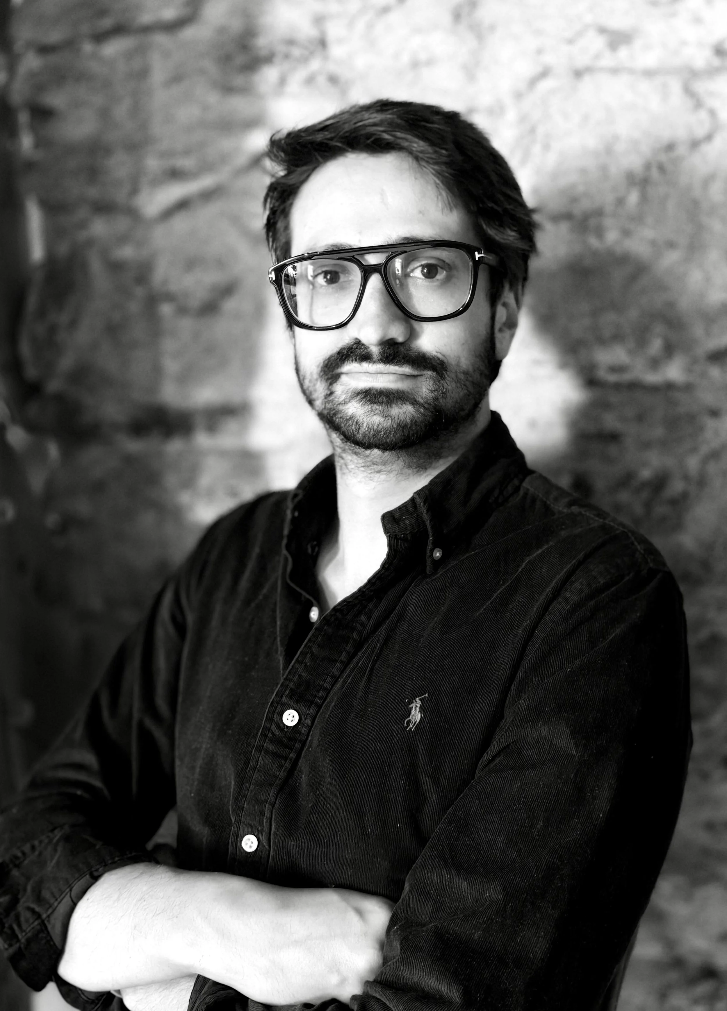 Homme portant des lunettes, moustache et barbe, dans une chemise noire avec un fond de mur en pierre.