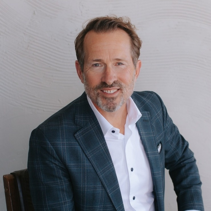 A middle-aged man with brown hair and a beard, wearing a dark plaid blazer and white shirt, sitting and smiling at the camera against a textured light gray wall.