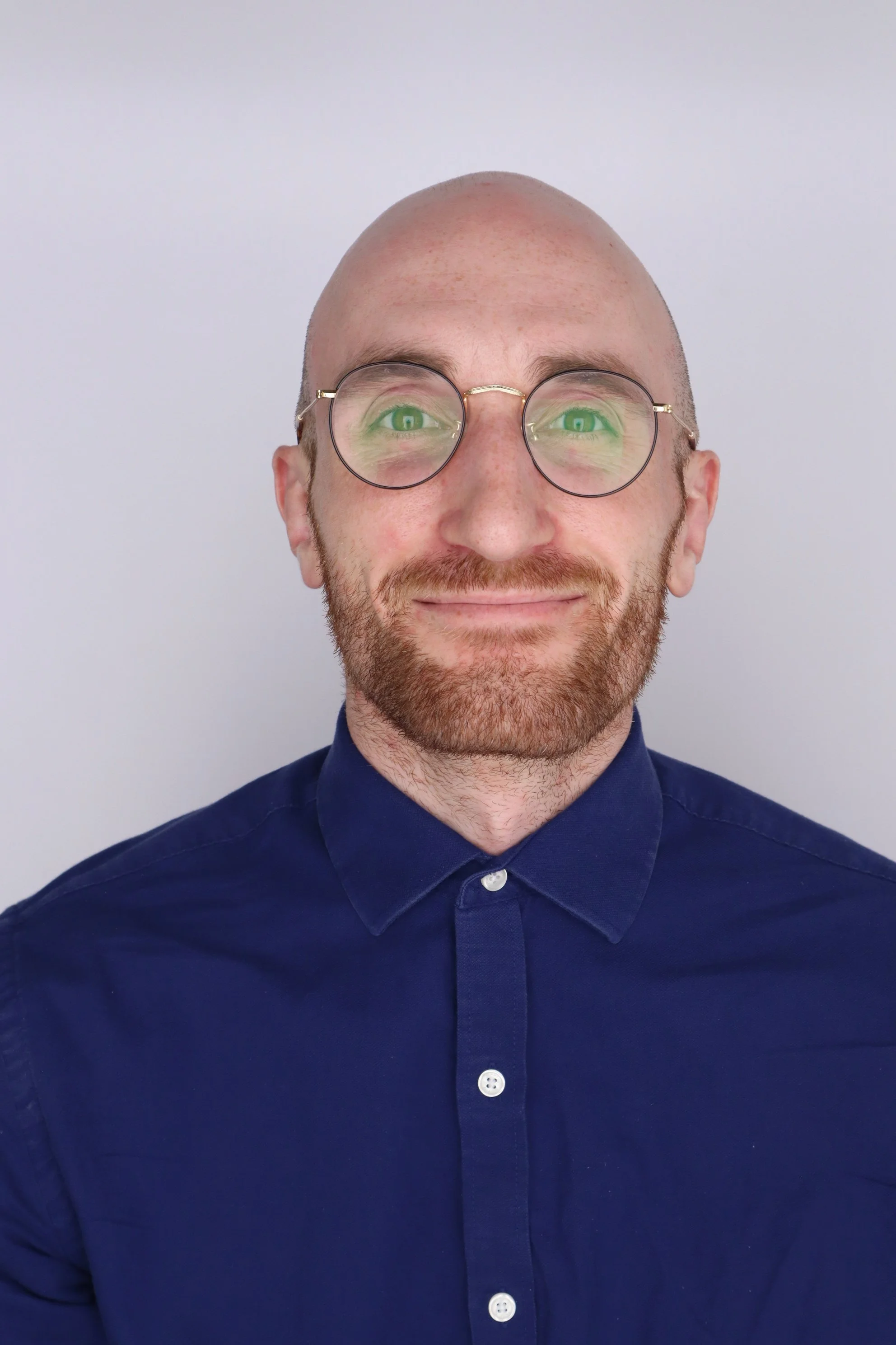 Portrait of a smiling man with glasses and a beard, wearing a dark blue button-up shirt, against a plain light-colored background.