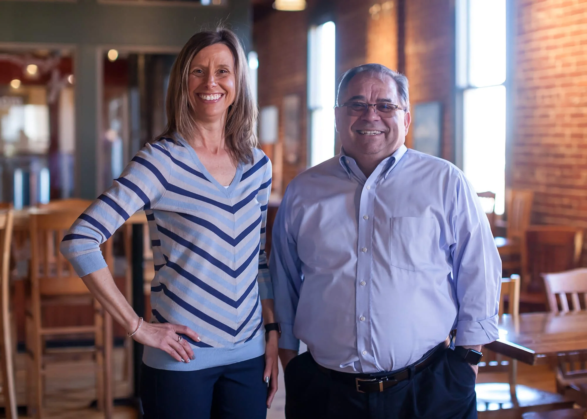 A woman and a man smiling in a restaurant with brick walls and large windows, dressed casually.