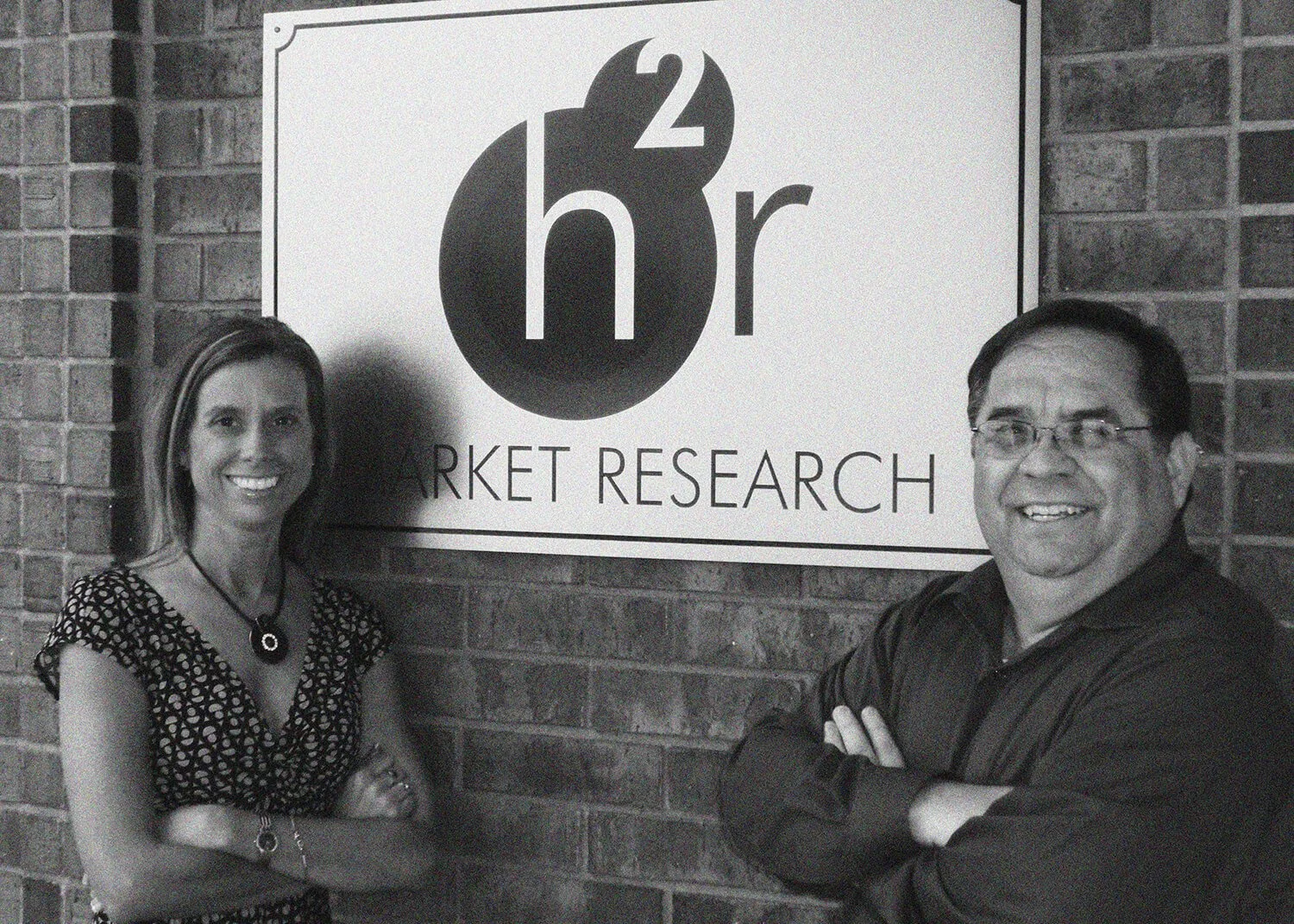 Two people, a woman with long hair and a man with glasses, smiling and standing with arms crossed in front of a sign that reads 'h²r Market Research' on a brick wall.