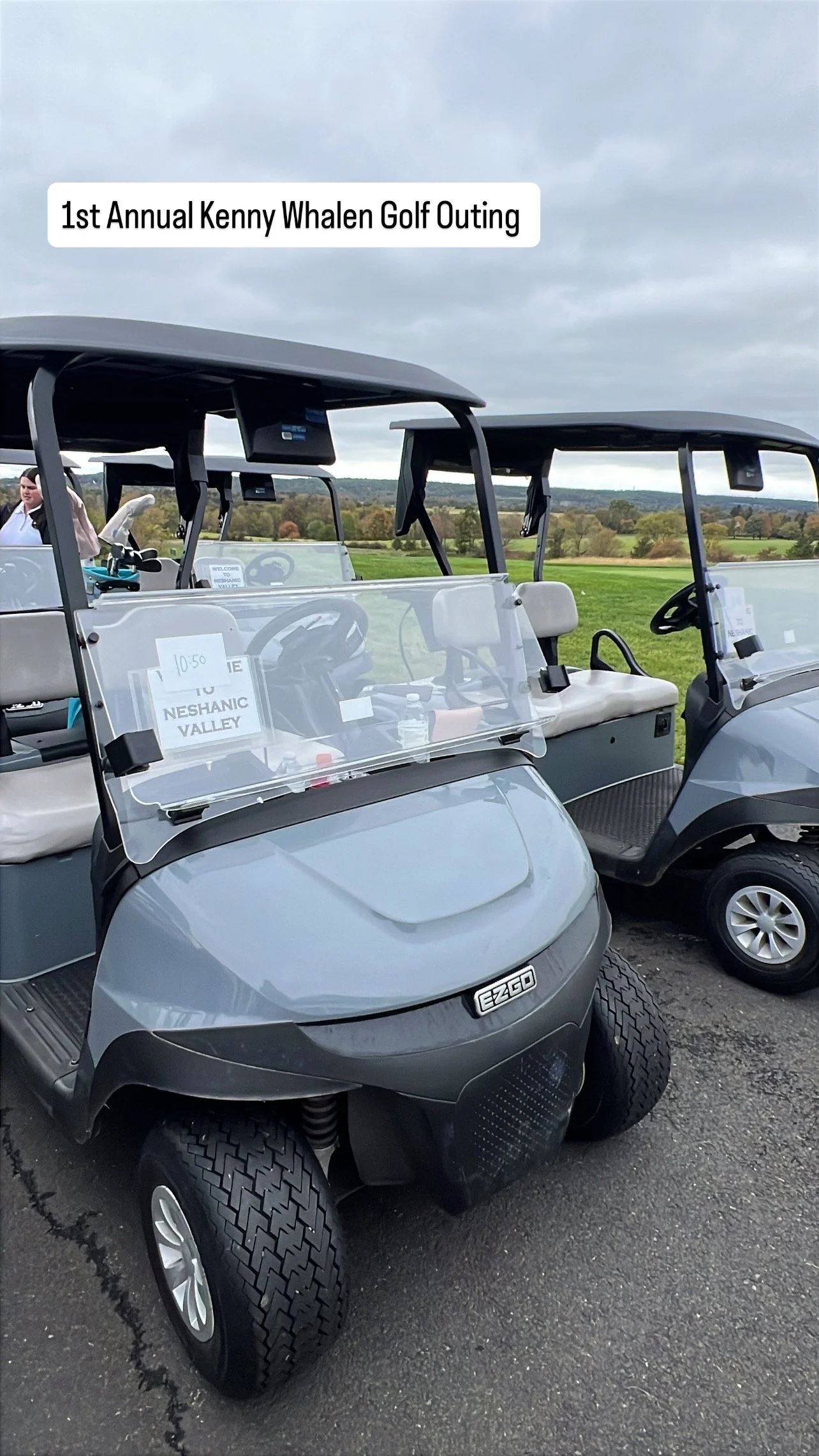 Two gray golf carts parked on a golf course with a cloudy sky and green landscape in the background, one cart with a sign that says '1st Annual Kenny Whalen Golf Outing'.