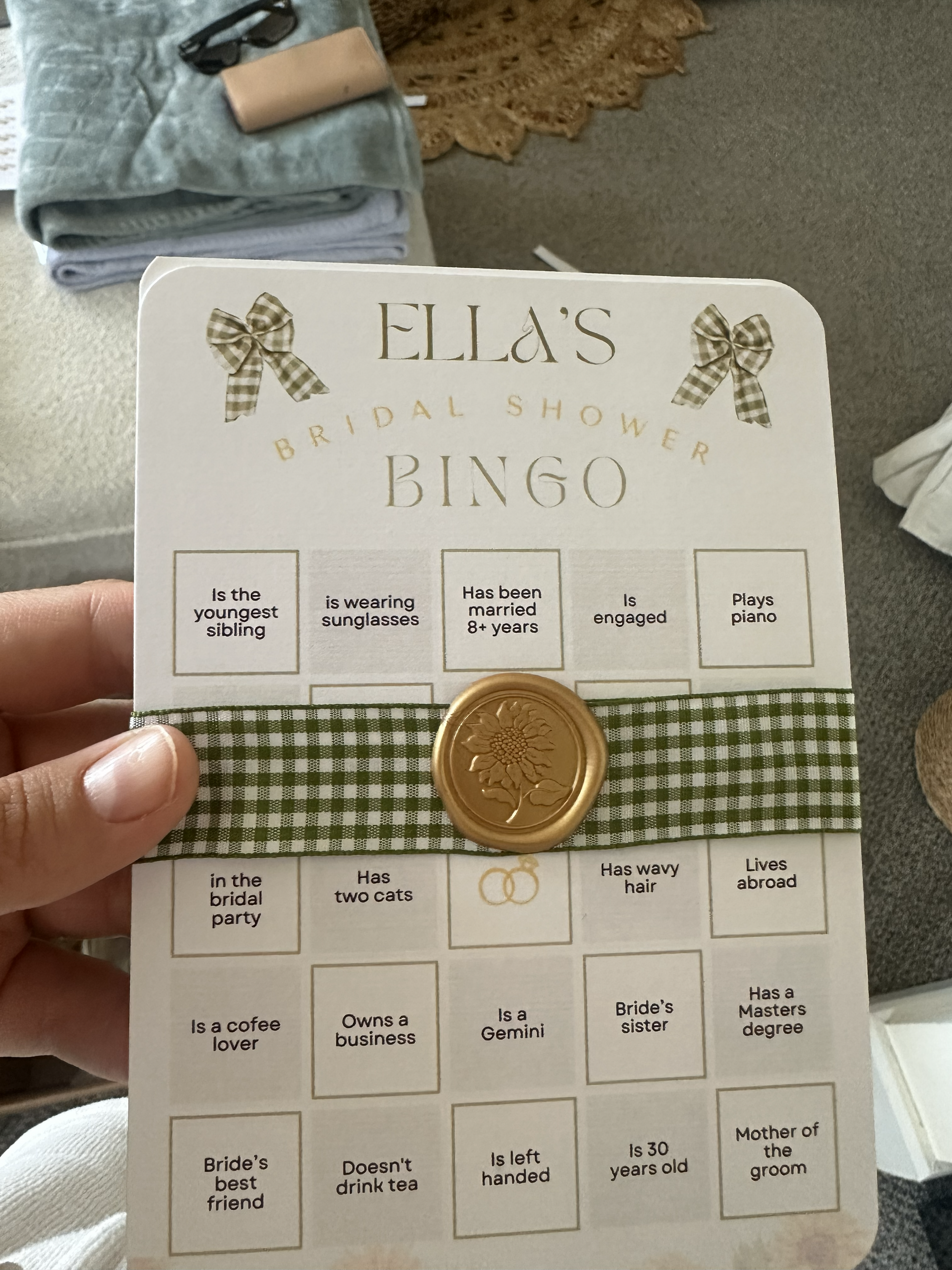 A bingo card with the title "Ella's Bridal Shower Bingo" featuring a checkered green and white ribbon and a gold wax seal with a flower design.