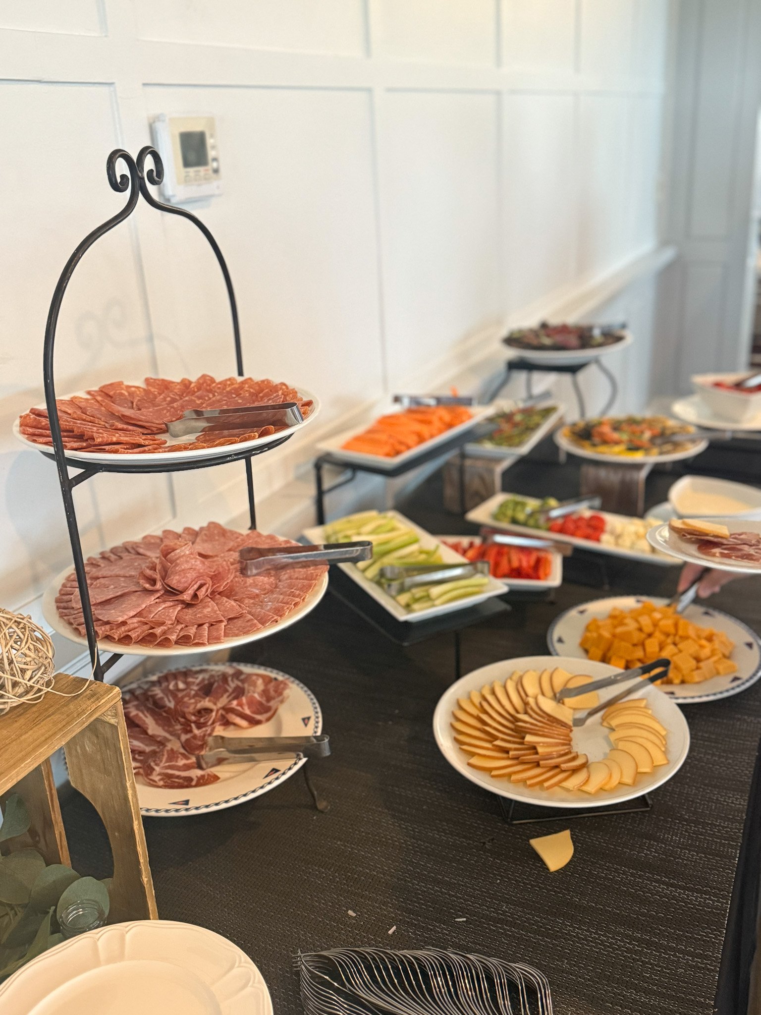 Buffet table with assorted meats, cheeses, vegetables, and garnishes on white plates and black trays.