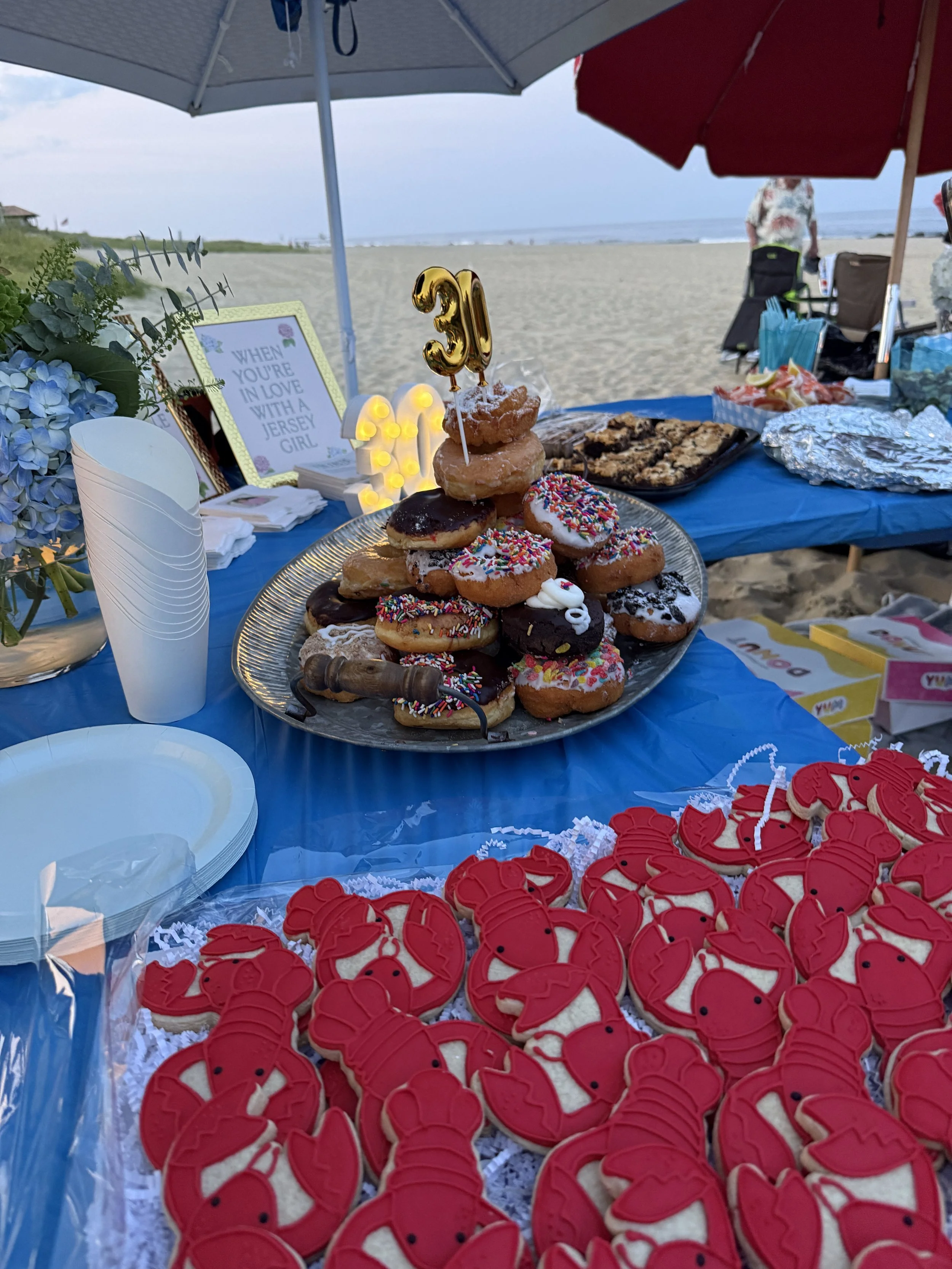 Cake and cookie desserts on a table at a beach party, with red lobster-shaped cookies and a layered ice cream treat topped with a gold number '31' candle. The table is decorated with a blue tablecloth and has a beach in the background with umbrellas 