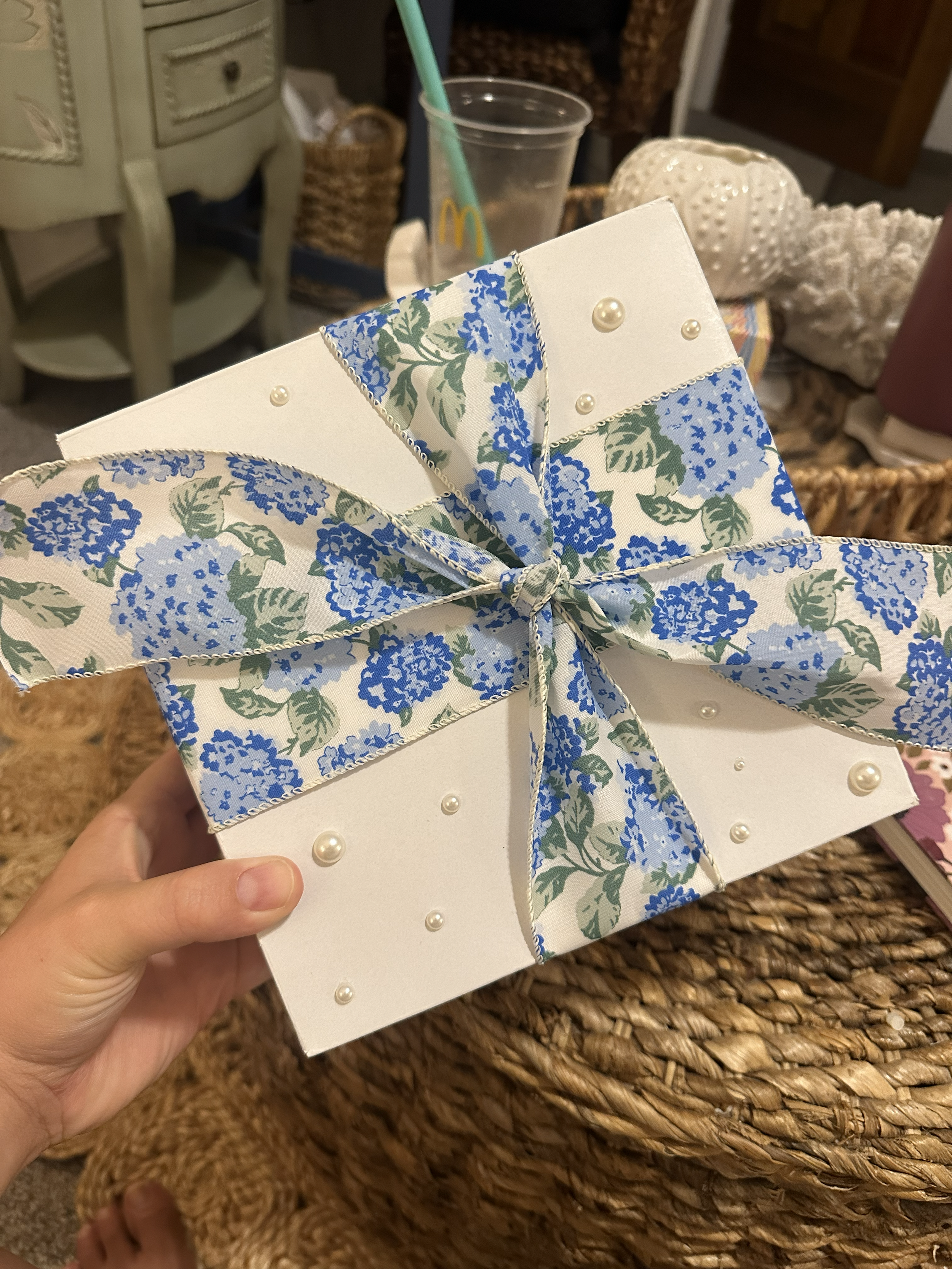 Hand holding a gift box decorated with a blue floral ribbon and pearl accents.