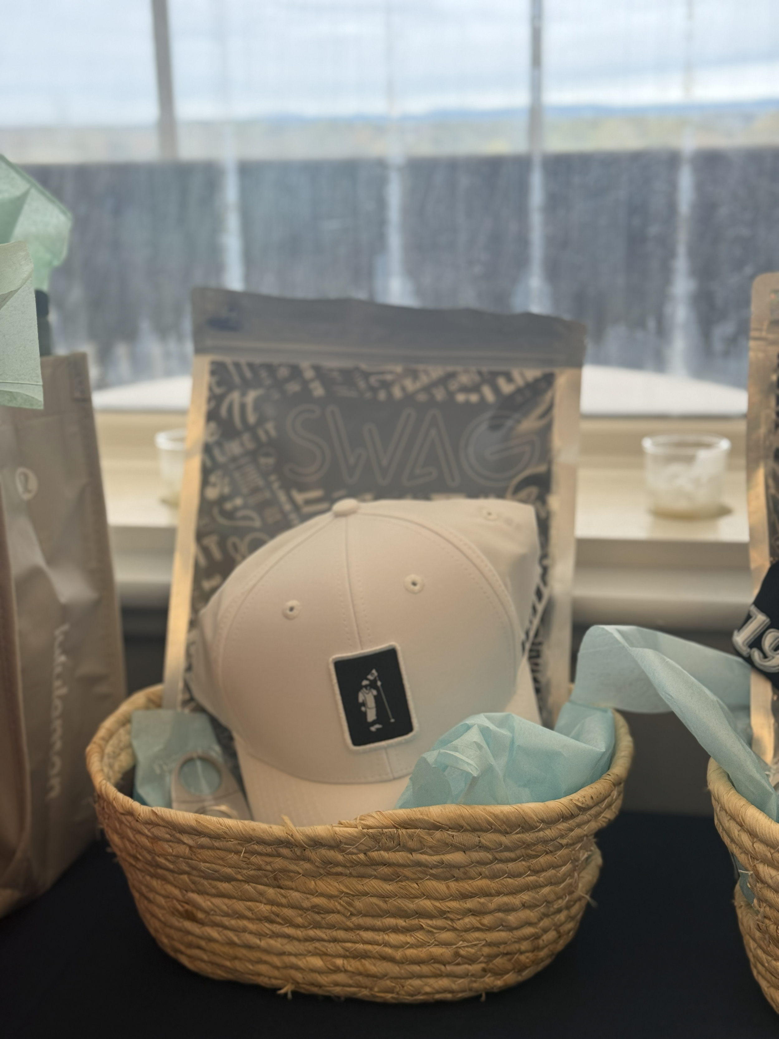 A white baseball cap with a golf-themed tag inside a woven basket with blue tissue paper, placed on a table with snack packages and a window in the background.