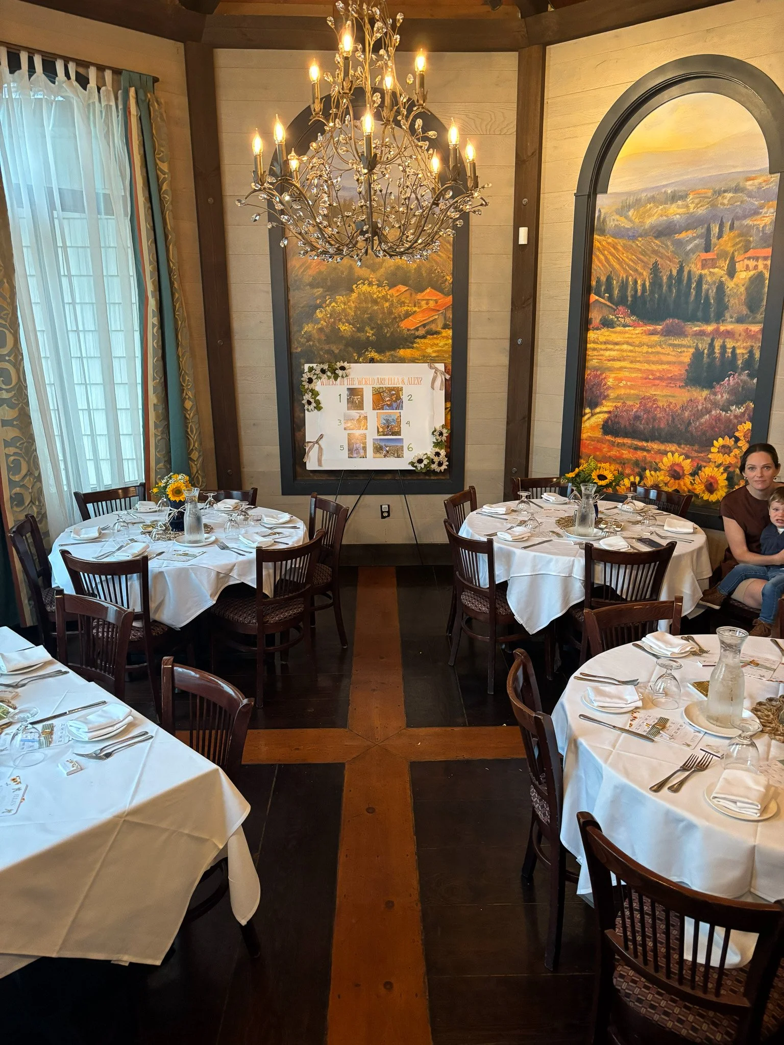 Elegantly set dining room with round tables covered in white tablecloths, decorated with sunflower centerpieces, crystal glassware, and silverware, illuminated by a large chandelier, with large landscape paintings on the walls.