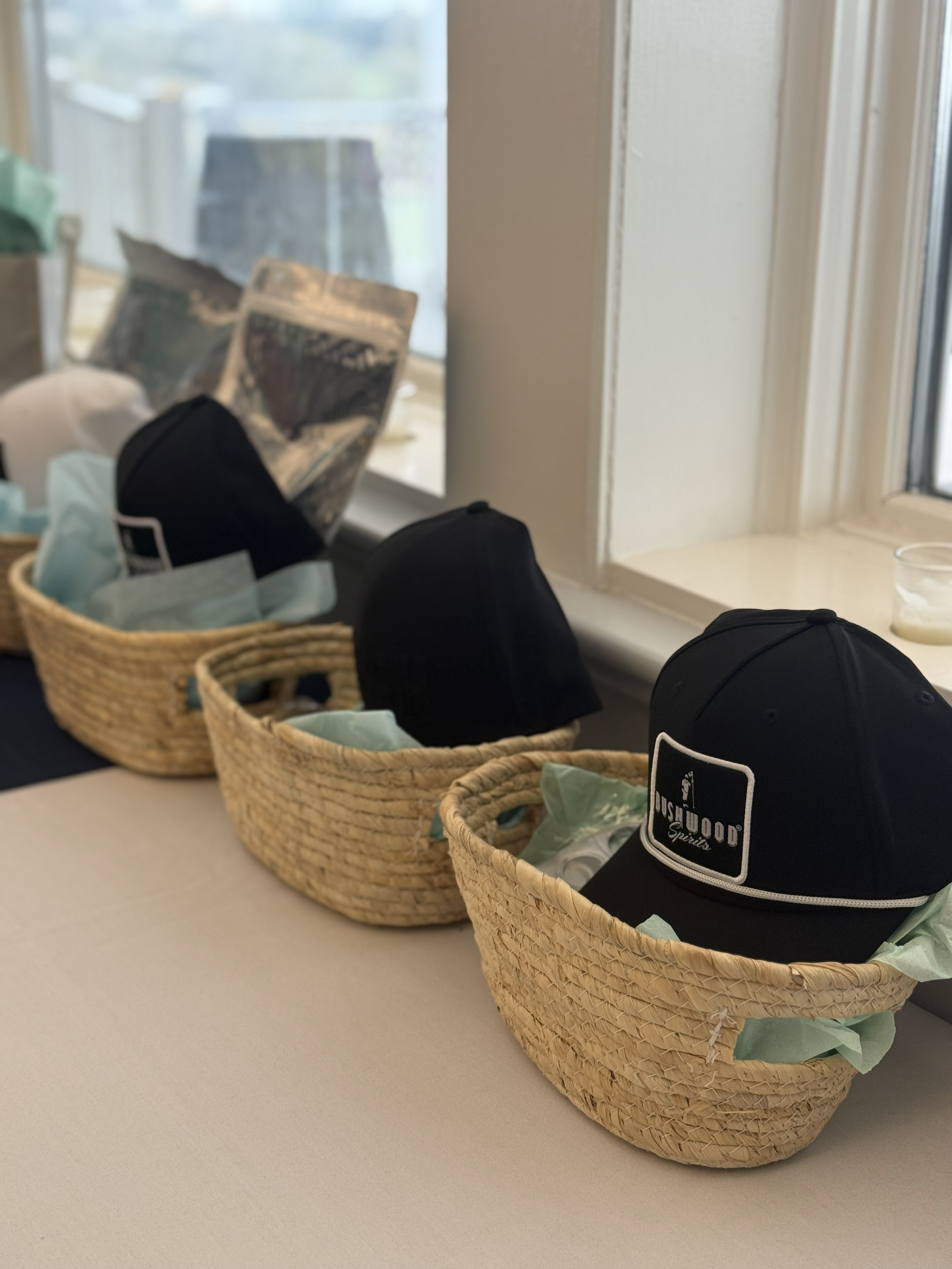 Three woven baskets containing black Caps with a logo, placed on a table near a window, with some tissue paper inside the baskets.