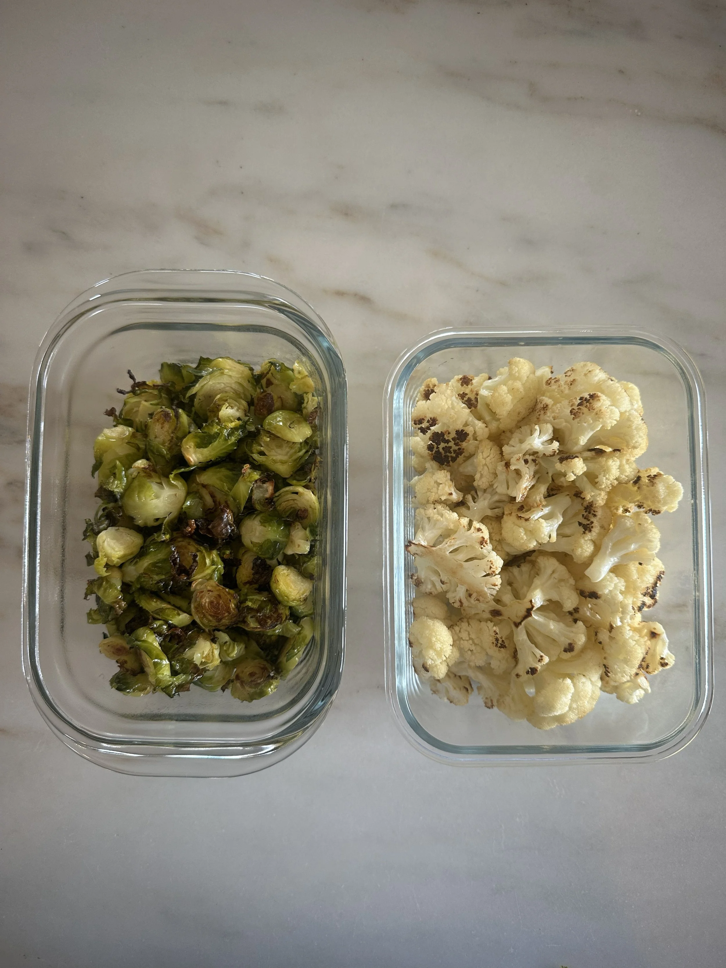 Roasted Brussels sprouts on the left and roasted cauliflower on the right in glass containers on a white countertop.