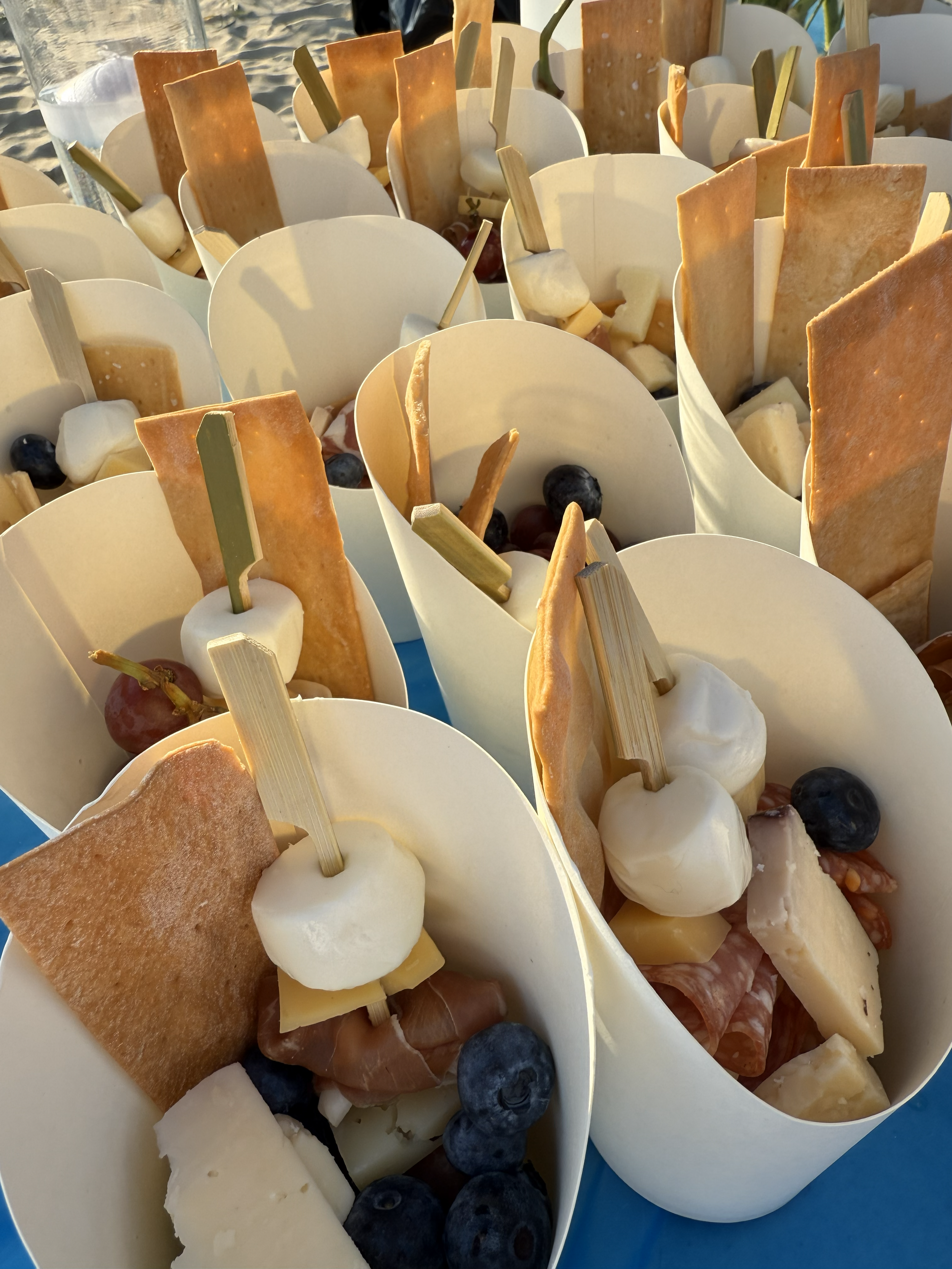 Multiple cups filled with cheese, grapes, cured meats, crackers, mozzarella balls, blueberries, and marshmallows, arranged outdoors.