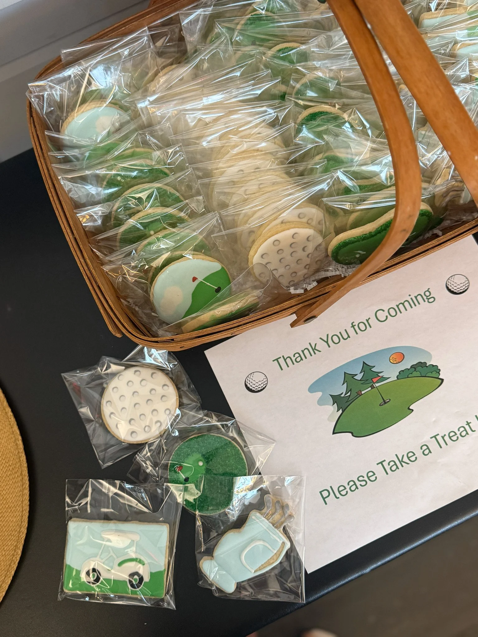 Variety of golf-themed cookies individually wrapped in plastic, placed in a basket and on a table. The cookies are decorated with golf course and golf club designs, along with a golf ball, and a golf tee.