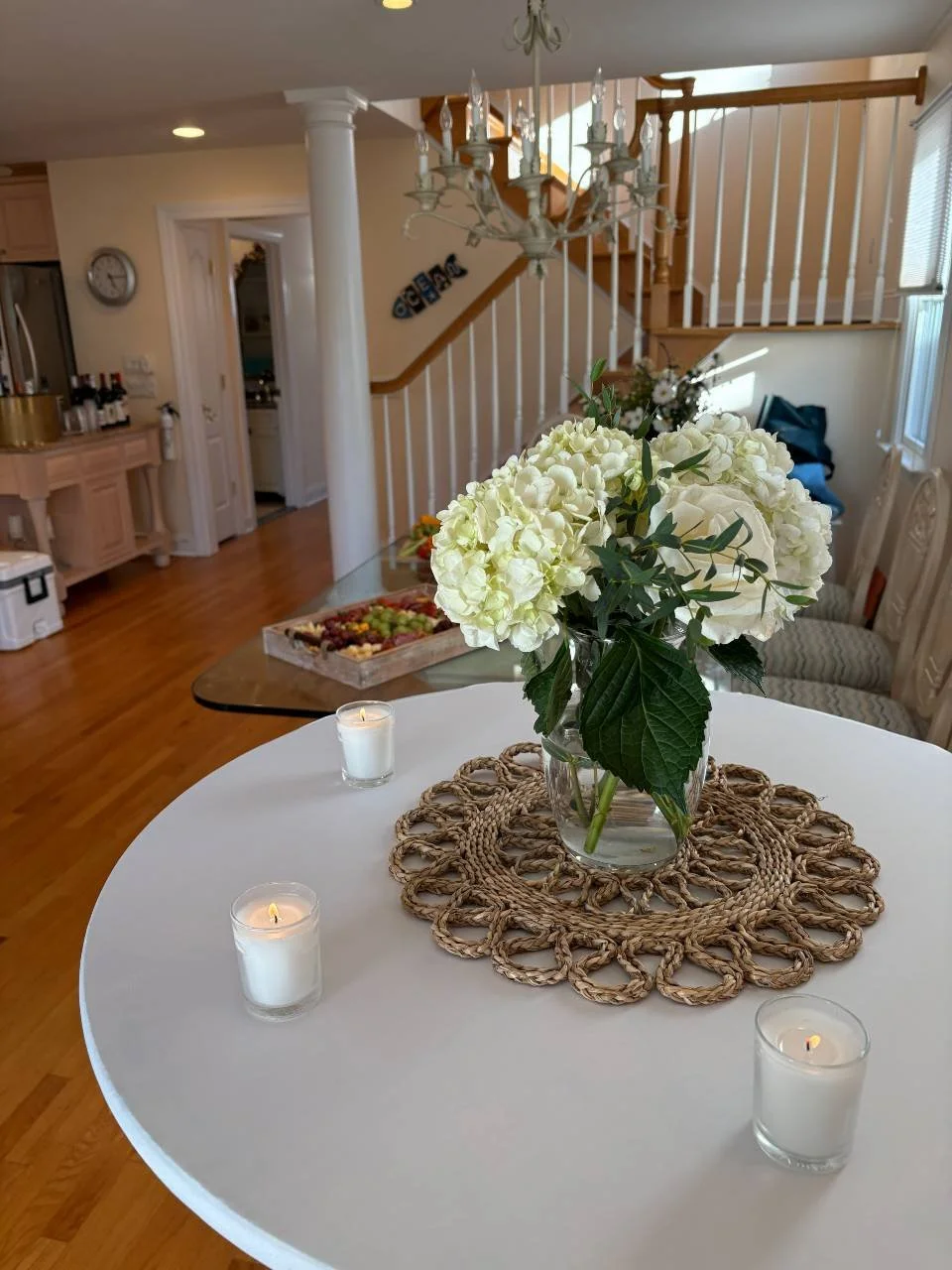 A white round table with a glass vase of white flowers, three lit white candles, and a woven placemat, inside a home with wooden floors, a staircase, and kitchen area visible in the background.
