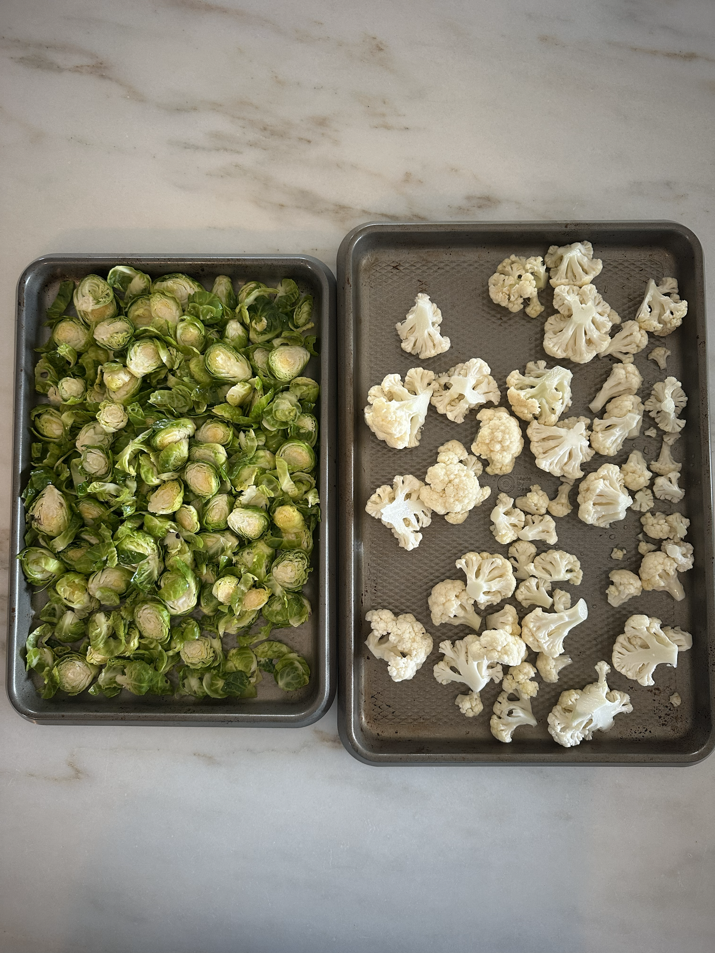 Two trays of vegetables on a marble countertop, one with Brussels sprouts and the other with cauliflower florets.