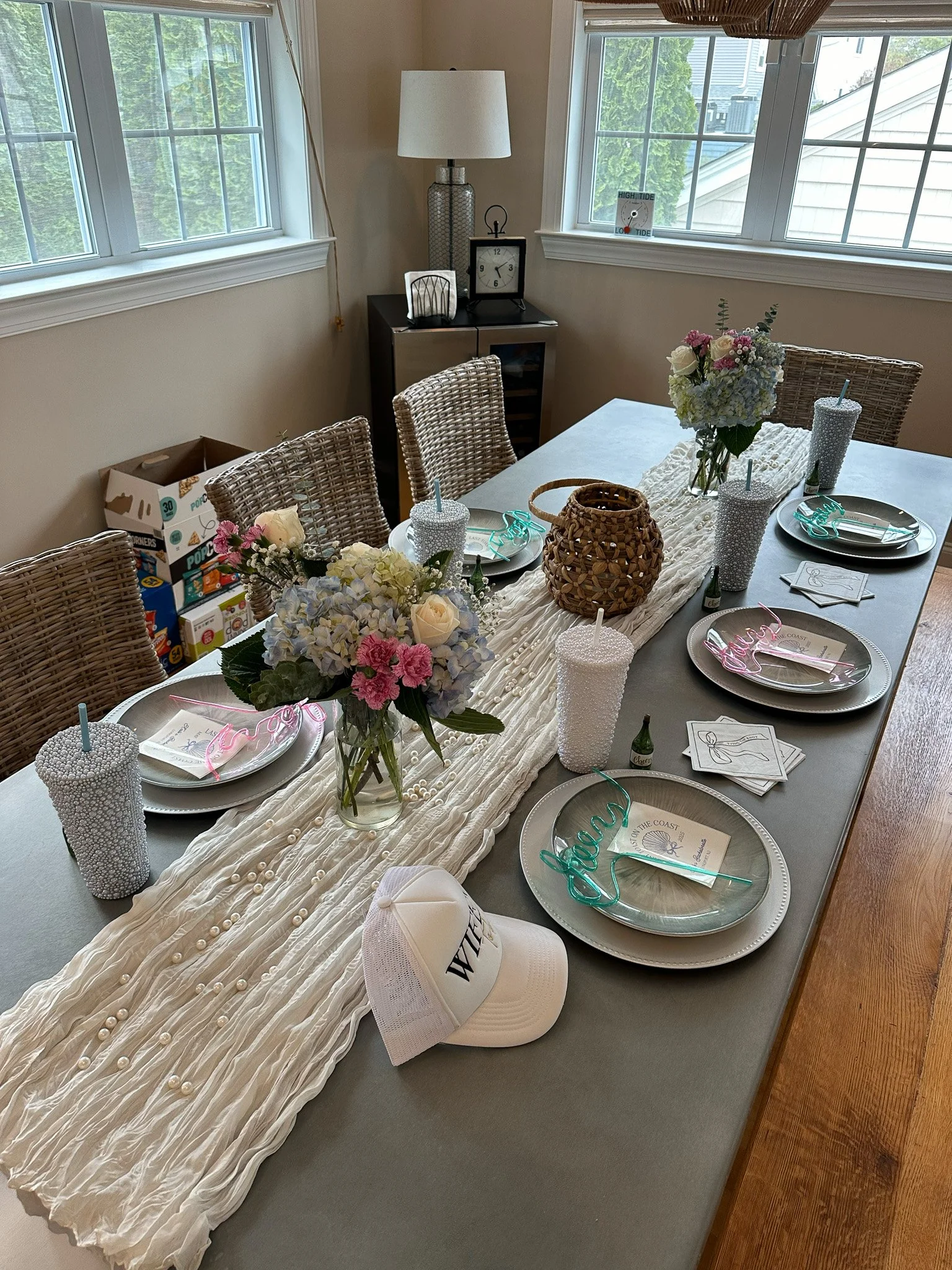 A dining table decorated for a celebration with a white, pleated table runner, vases of flowers, pearl-covered cups, and customized stir sticks. The table has place settings with plates, napkins, and glasses, and a tan woven basket. In the background