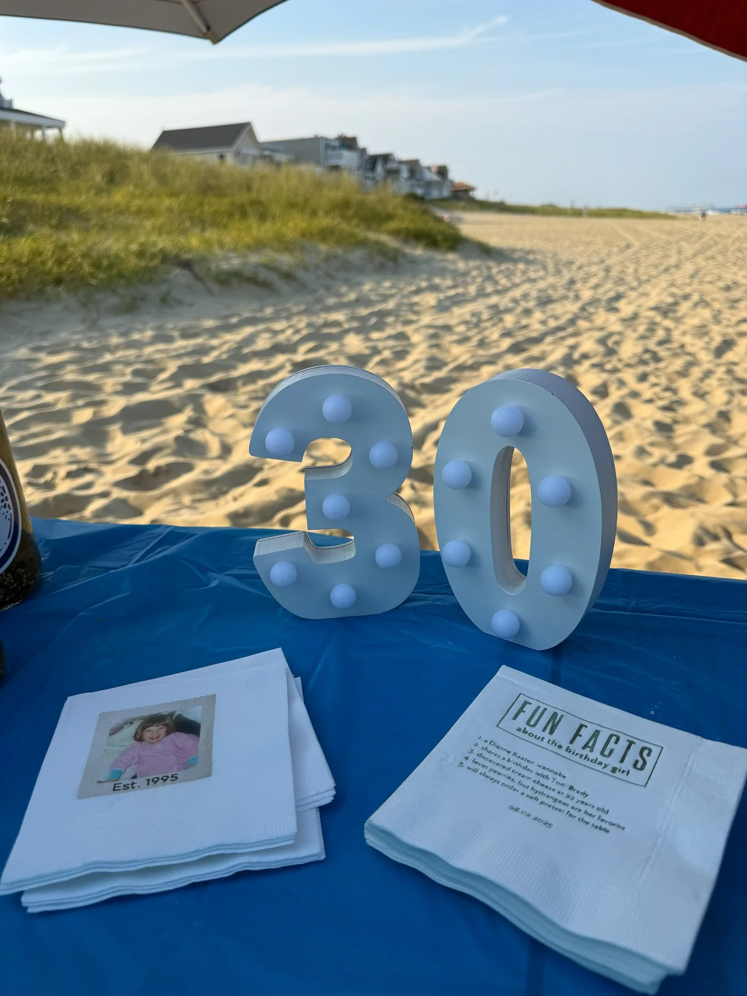 Beach scene with a large white illuminated number '30' on a blue table, photographs, and fun facts sheet, with sandy beach, grassy dunes, houses, and a cloudy sky in the background.