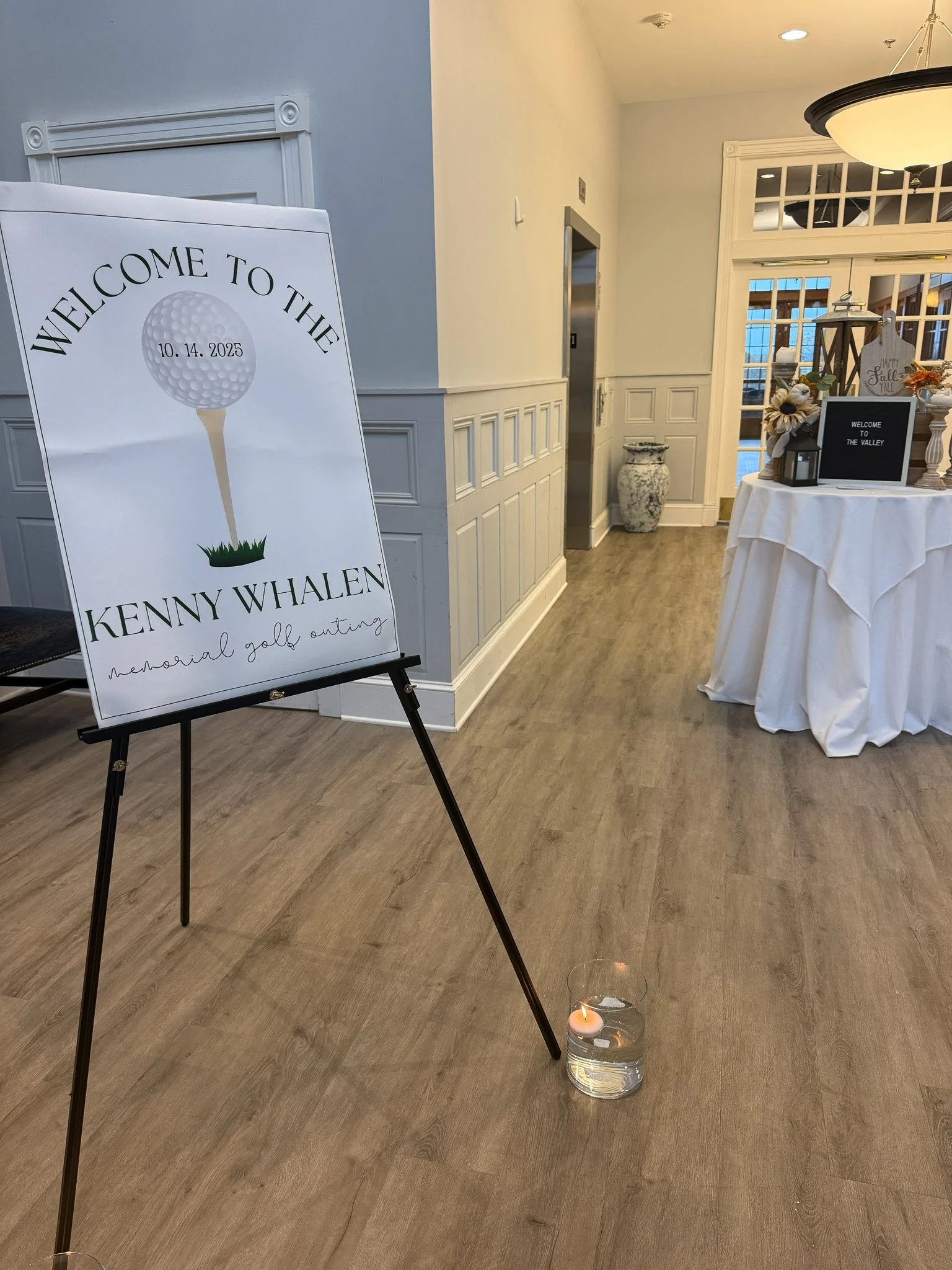 Event signboard with golf ball illustration and text welcoming attendees to Kenny Whalen's memorial golf outing, placed on an easel inside a well-lit room with wooden flooring and decorated table.