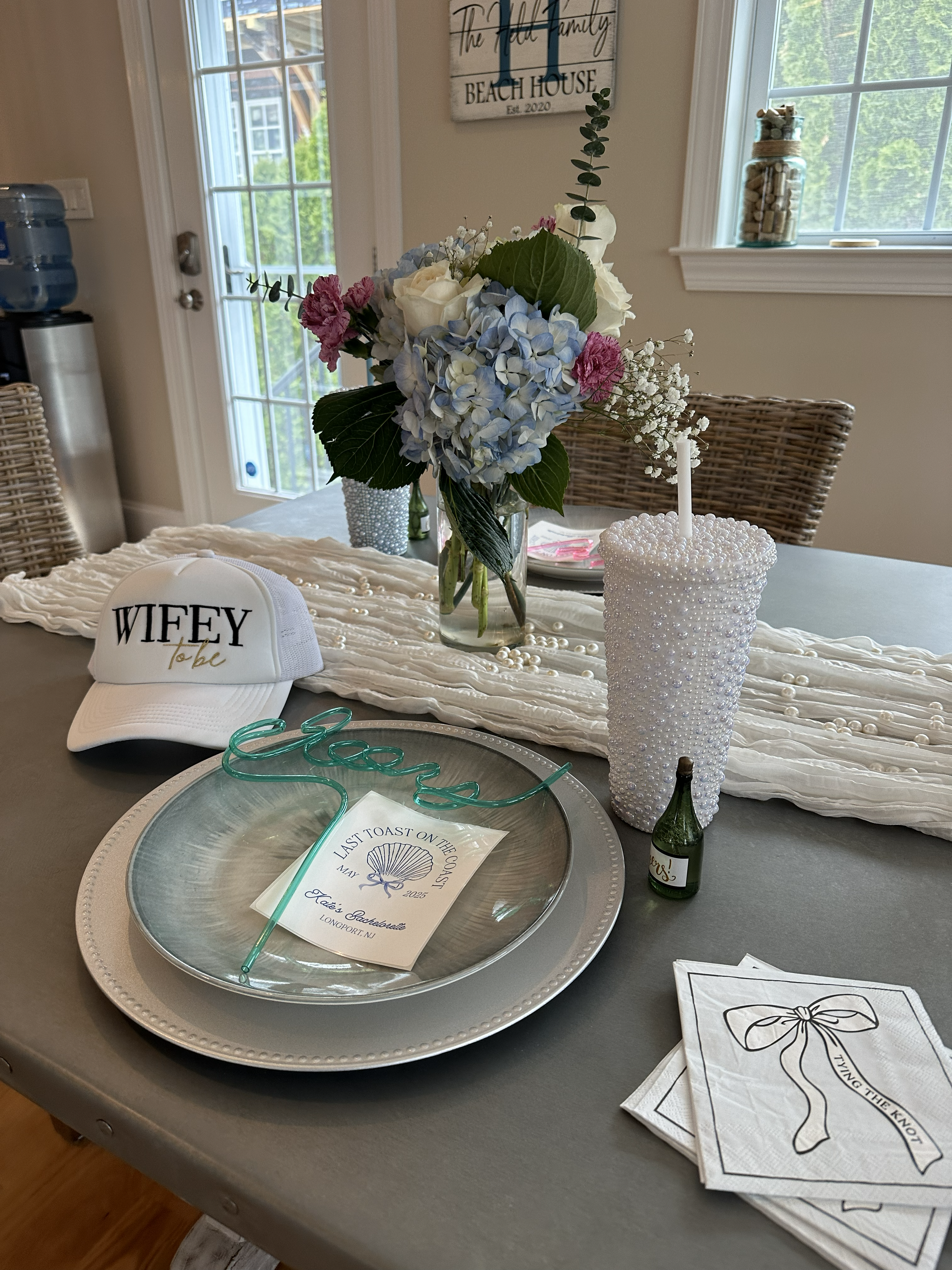 Decorative table setting with a large bouquet of white, pink, and blue flowers in a glass vase, a white giggler hat with 'Wifey to be' written on it, a large beaded cup with a straw, a small green drink bottle, a silver tray with a seashell card that