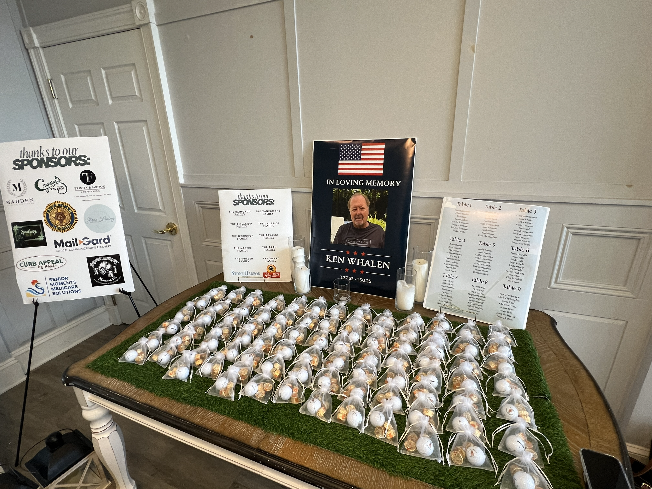 Memorial display with candles, and small personalized favors on a green felt runner on a wooden table, set against a beige paneled wall. Signs showing sponsors and a seating chart are nearby.