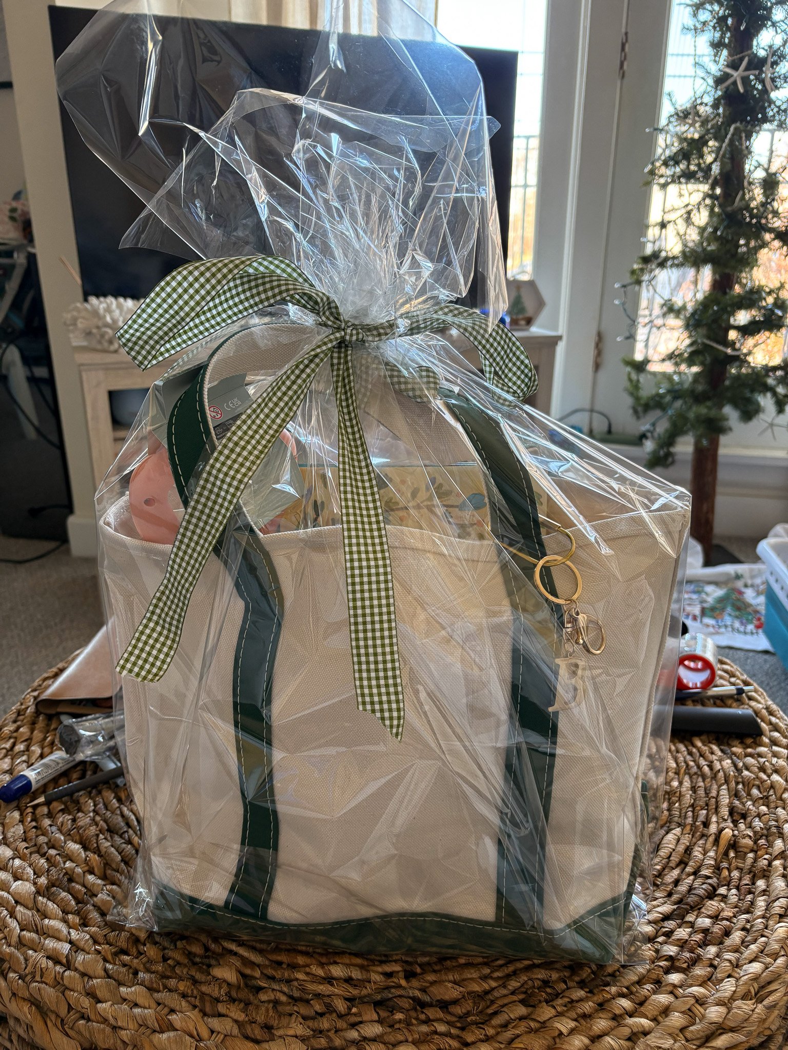 Gift bag wrapped in clear plastic with green and white checkered ribbon, containing a tote bag and other items, placed on a woven table.