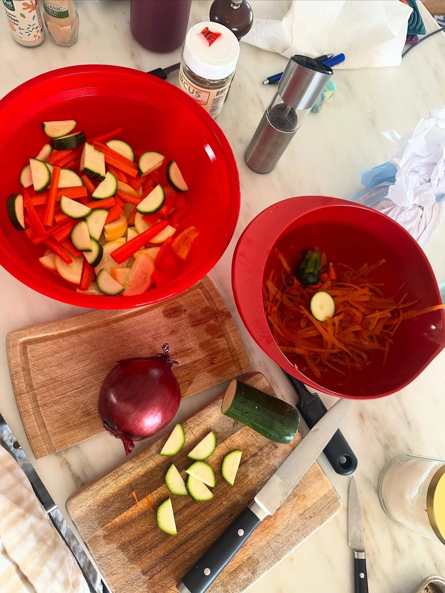 A kitchen countertop with chopped vegetables including zucchini, onion, and red bell pepper in red bowls, a knife, a chopping board, and various spice bottles.