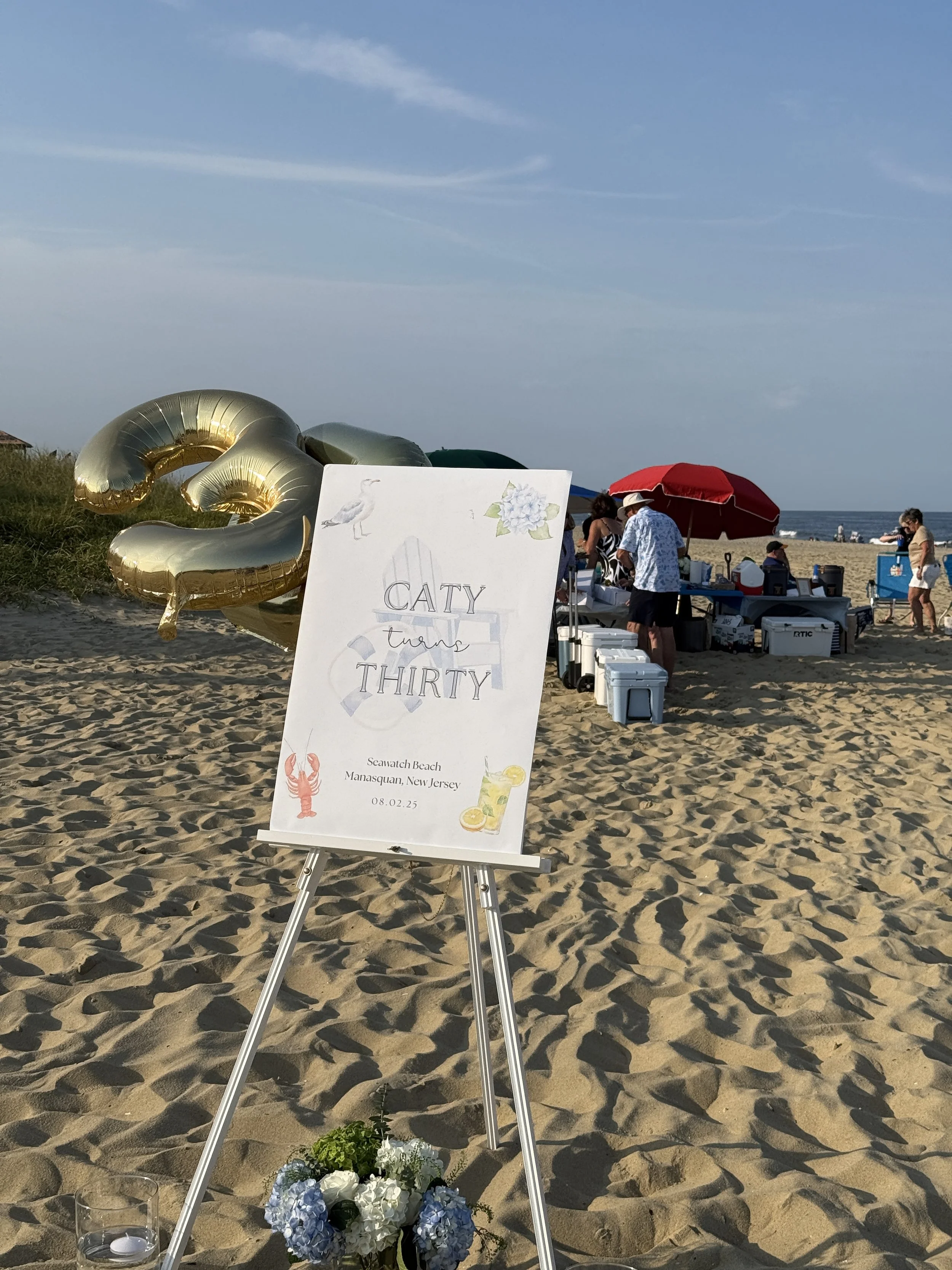 A beach scene with a white sign celebrating a 30th birthday for Caty, featuring blue and white flowers, a lobster, and a drink illustration. Gold balloons and a seagull graphic are attached, with beachgoers, coolers, and umbrellas in the background.