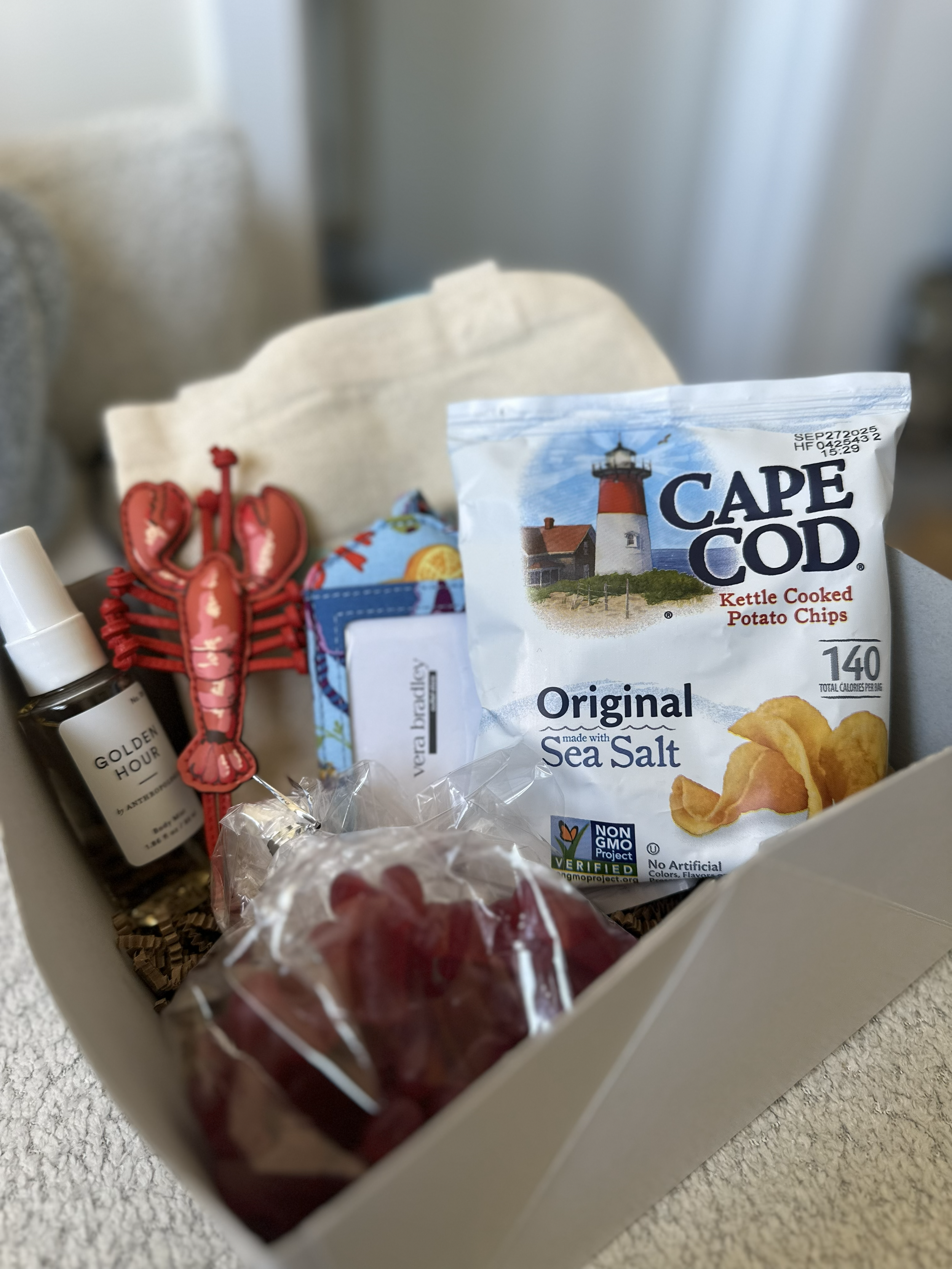 A gift box containing a miniature lobster bottle, a small bag of red grapes, a bag of Cape Cod Original Sea Salt Kettle Cooked Potato Chips, and a white cloth item, possibly a napkin or towel.