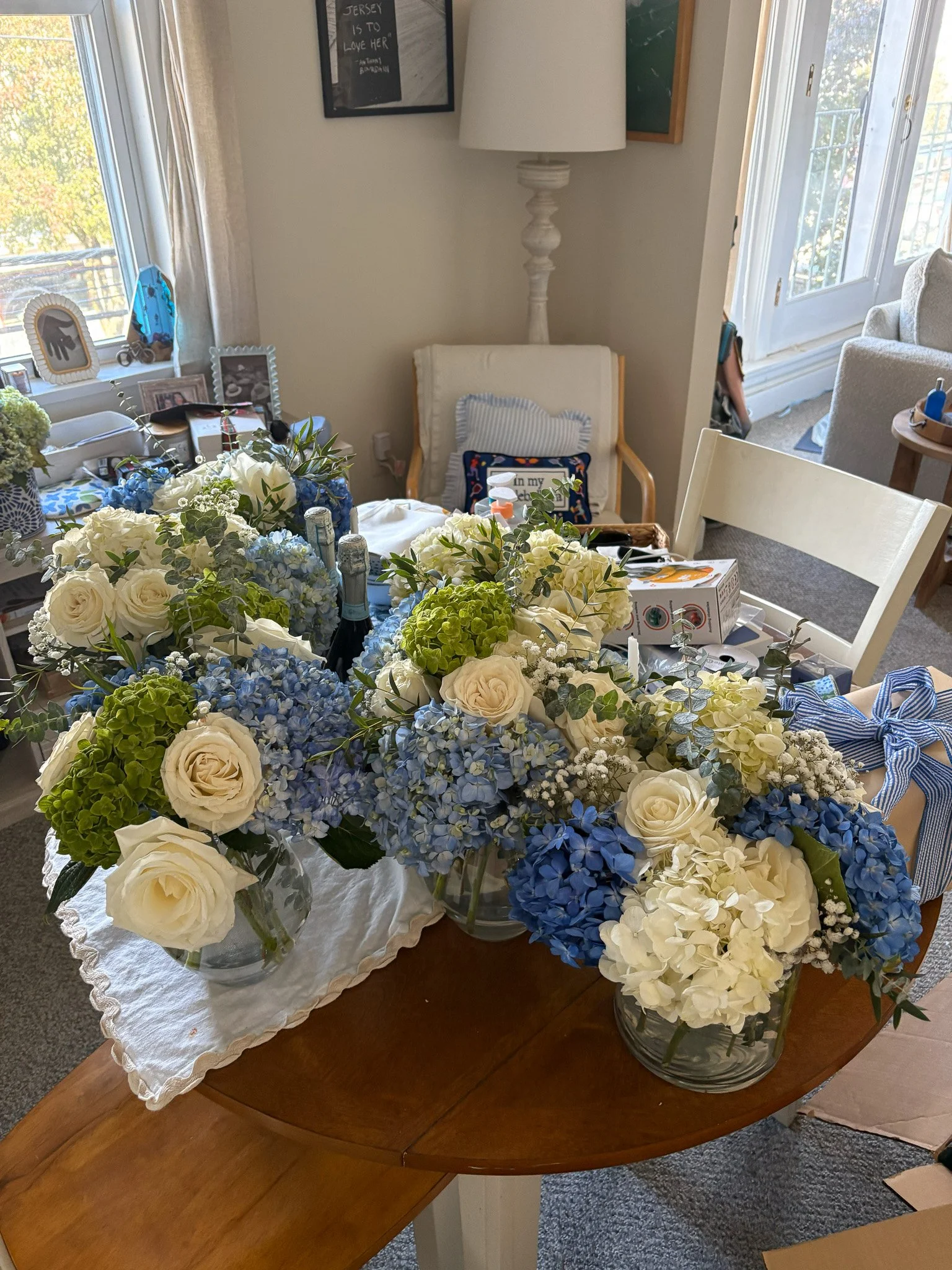 A wooden dining table with multiple glass vases filled with white and blue hydrangeas and white roses, set in a bright room with a window, framed pictures, and a white chair with pink and blue cushions in the background.