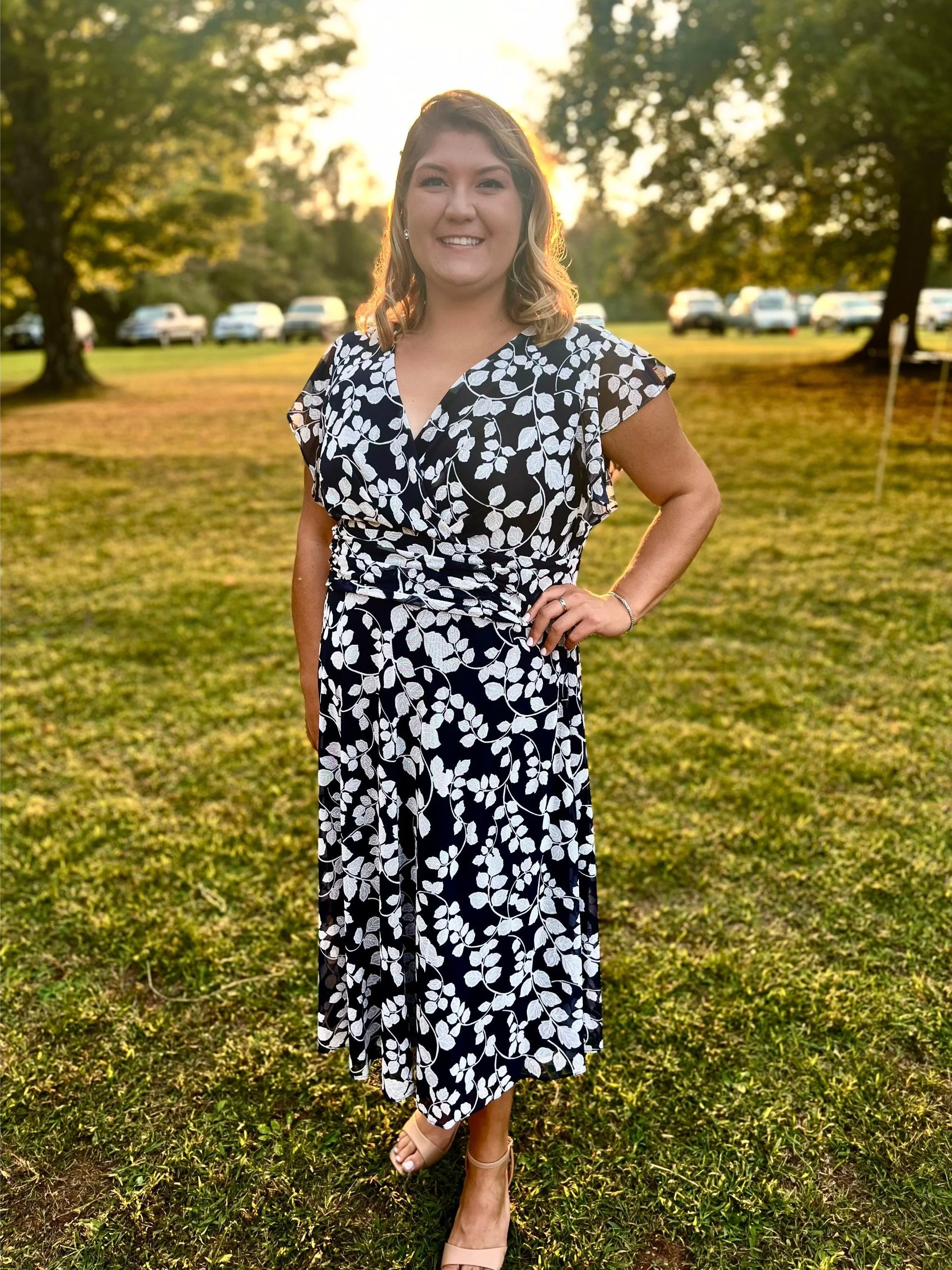 A smiling woman standing on grass in a black and white floral dress, with her hand on her hip, in a park during sunset.